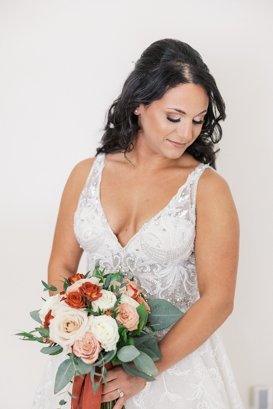 A bride smiles down her shoulder while standing in a white room holding her red and white rose bouquet