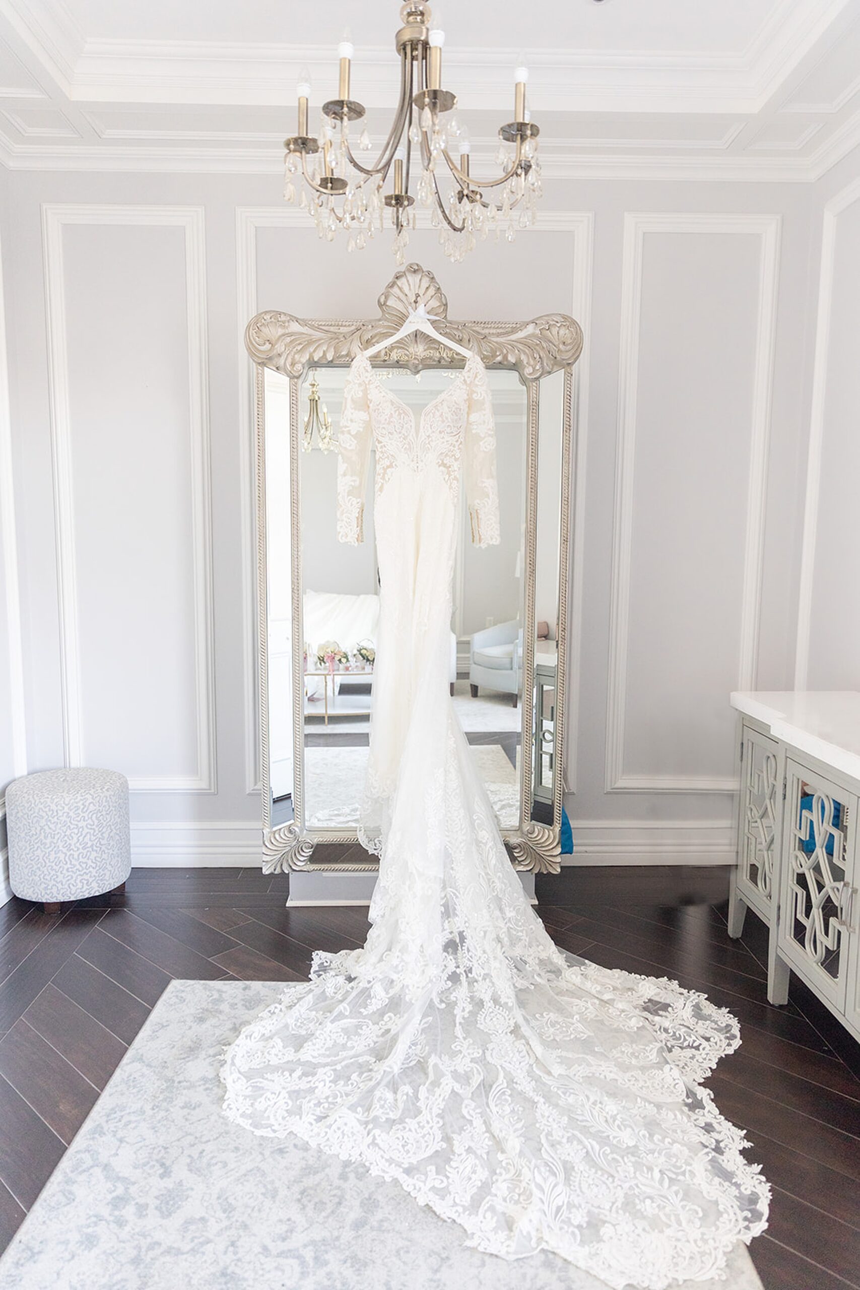 A wedding dress with a long lace train hangs from a large ornate mirror under a chandelier