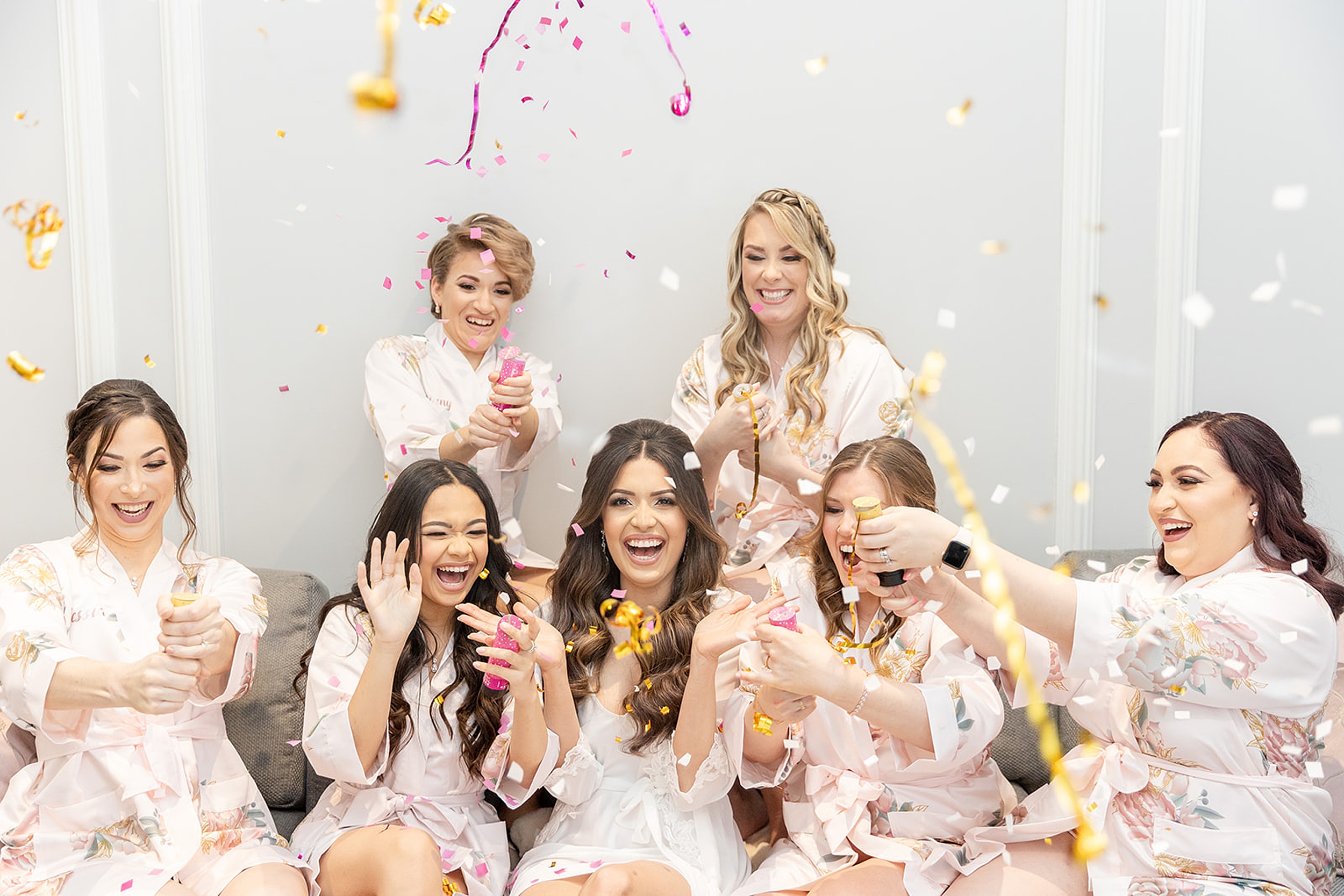 A bride sits on a couch with her bridesmaids as they pop confetti and streamers