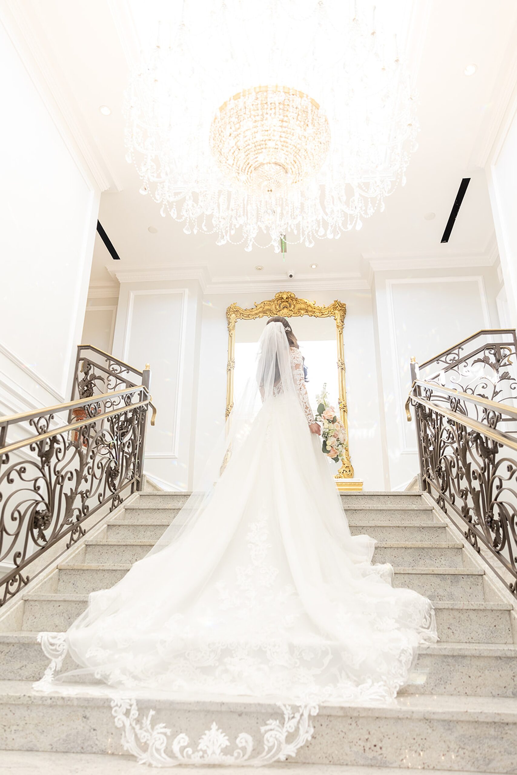 A bride looks up to a large crystal chandelier on a grand marble staircase