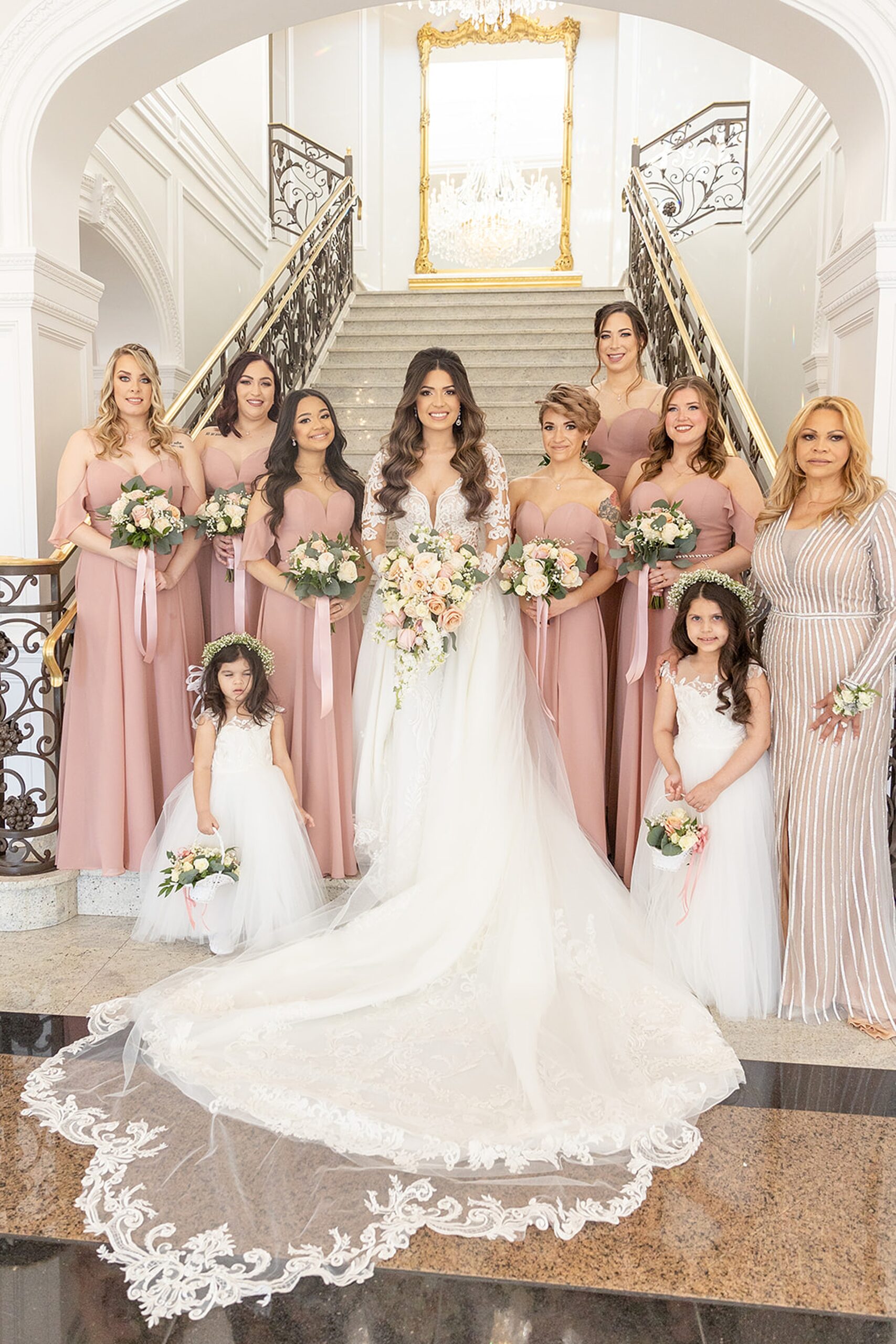 A bride in a long lace dress stands with her wedding party at the bottom of a grand marble staircase at the rockleigh wedding venue