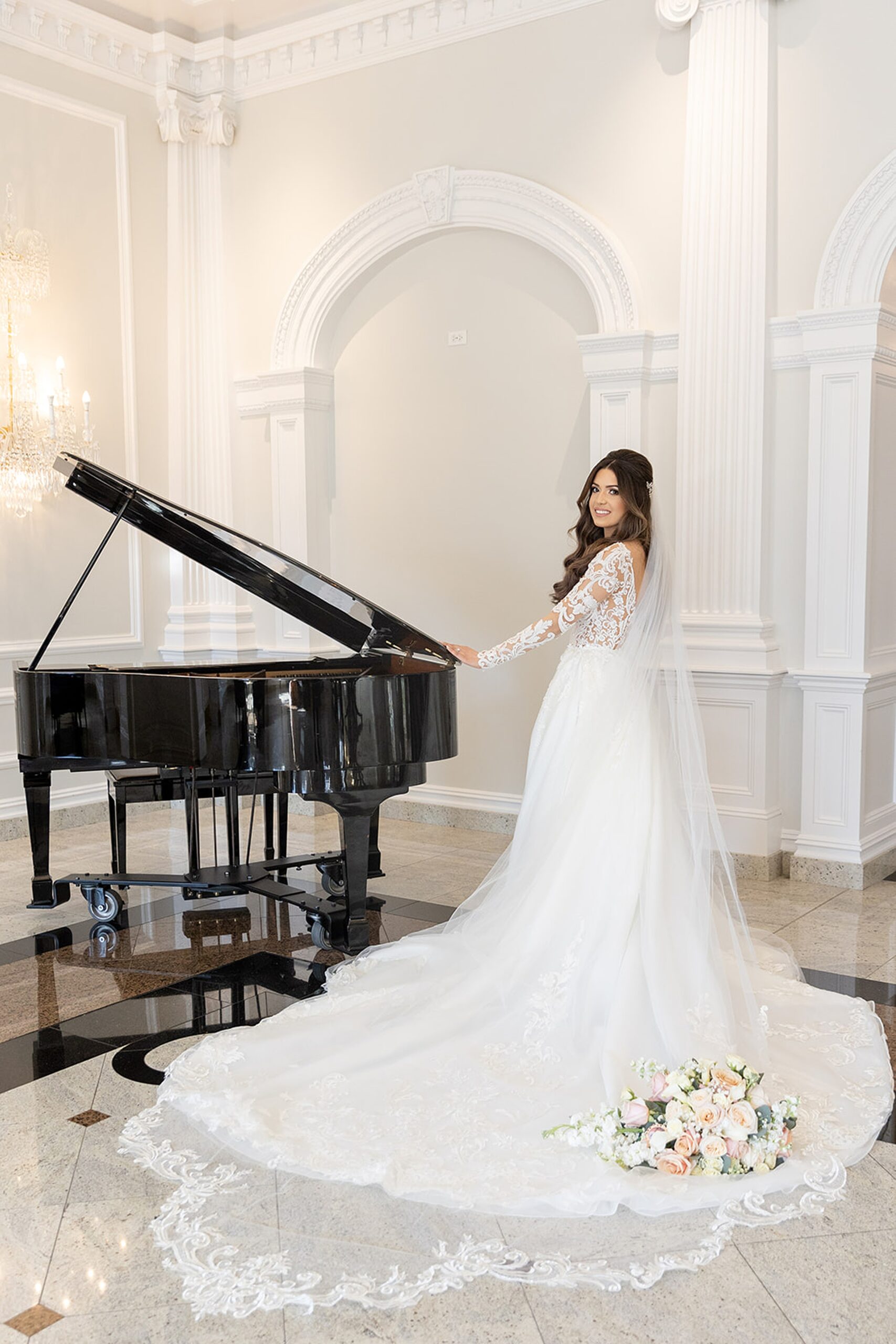A bride in a long flowing lace dress stands at a piano in a lobby with her bouquet laying in her train at the rockleigh wedding venue