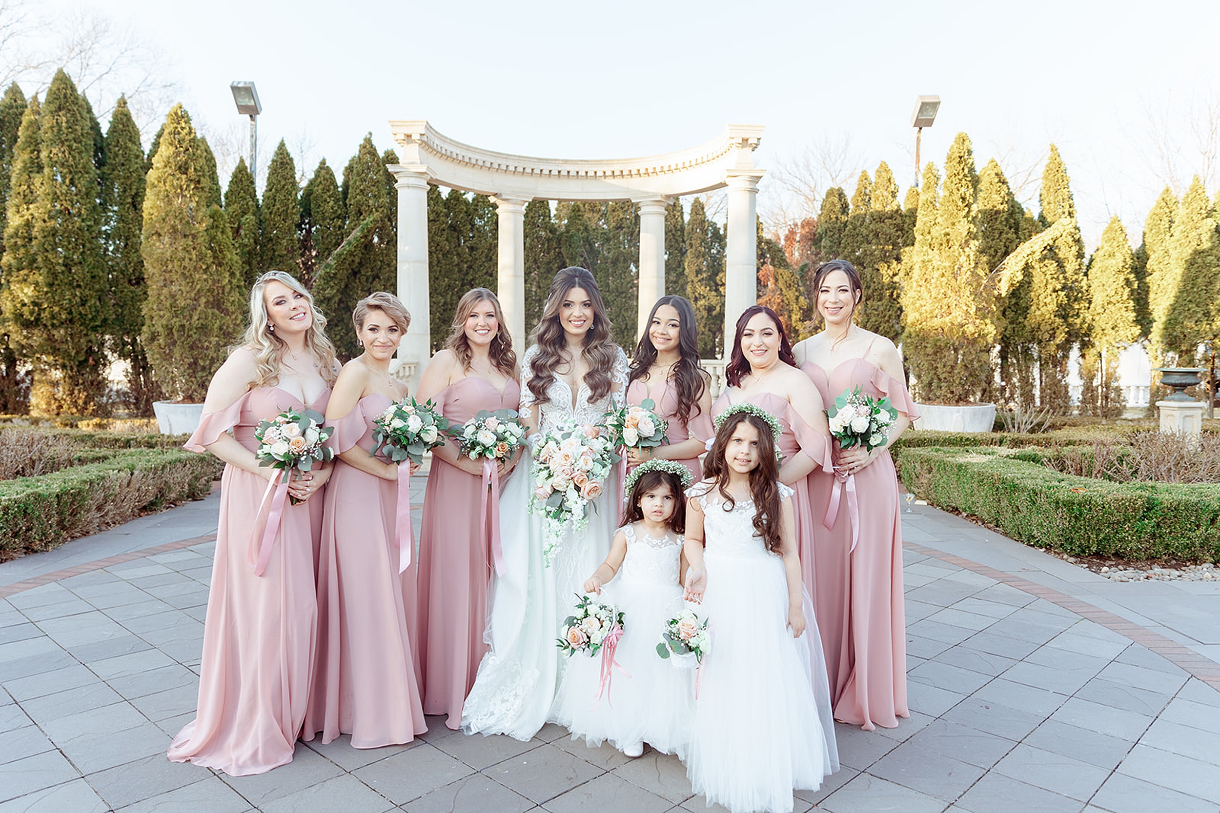 A bride stands with her bridal party and flower girls in a garden patio holding their bouquets