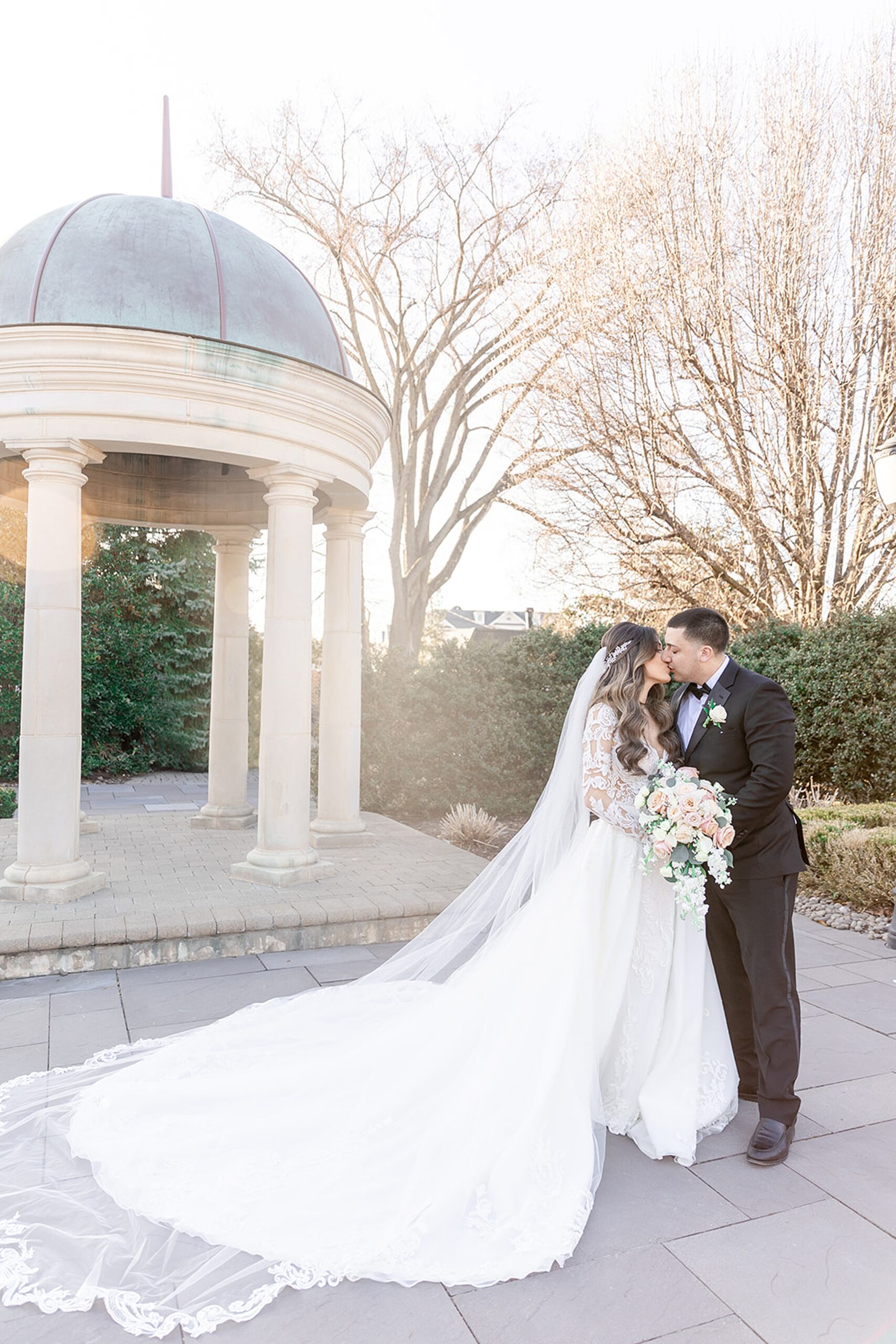 Newlyweds kiss with the long dress and veil flowing behind them at sunset by a copper top gazebo at the rockleigh wedding venue