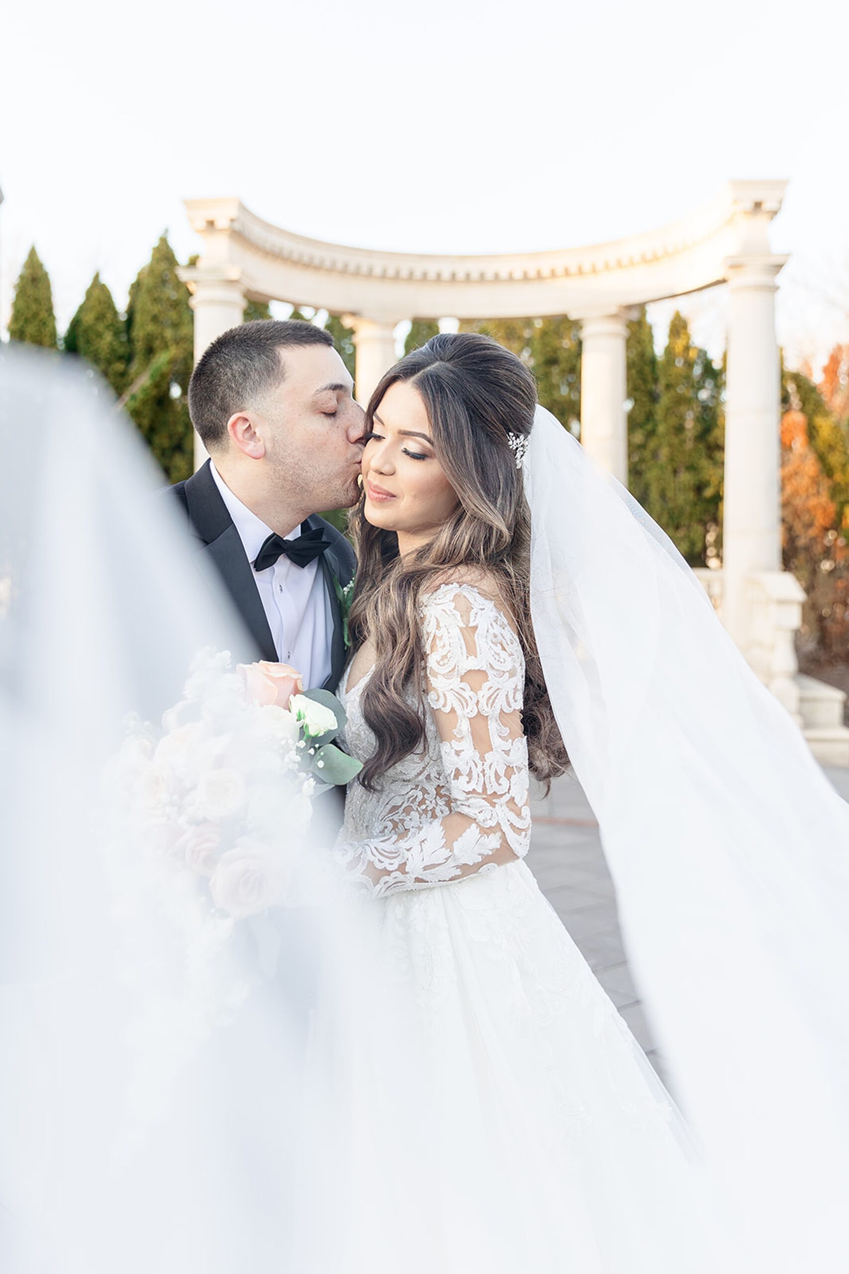 A groom in a black tuxedo kisses his bride in a white lace dress while the veil blows in the wind in a garden patio