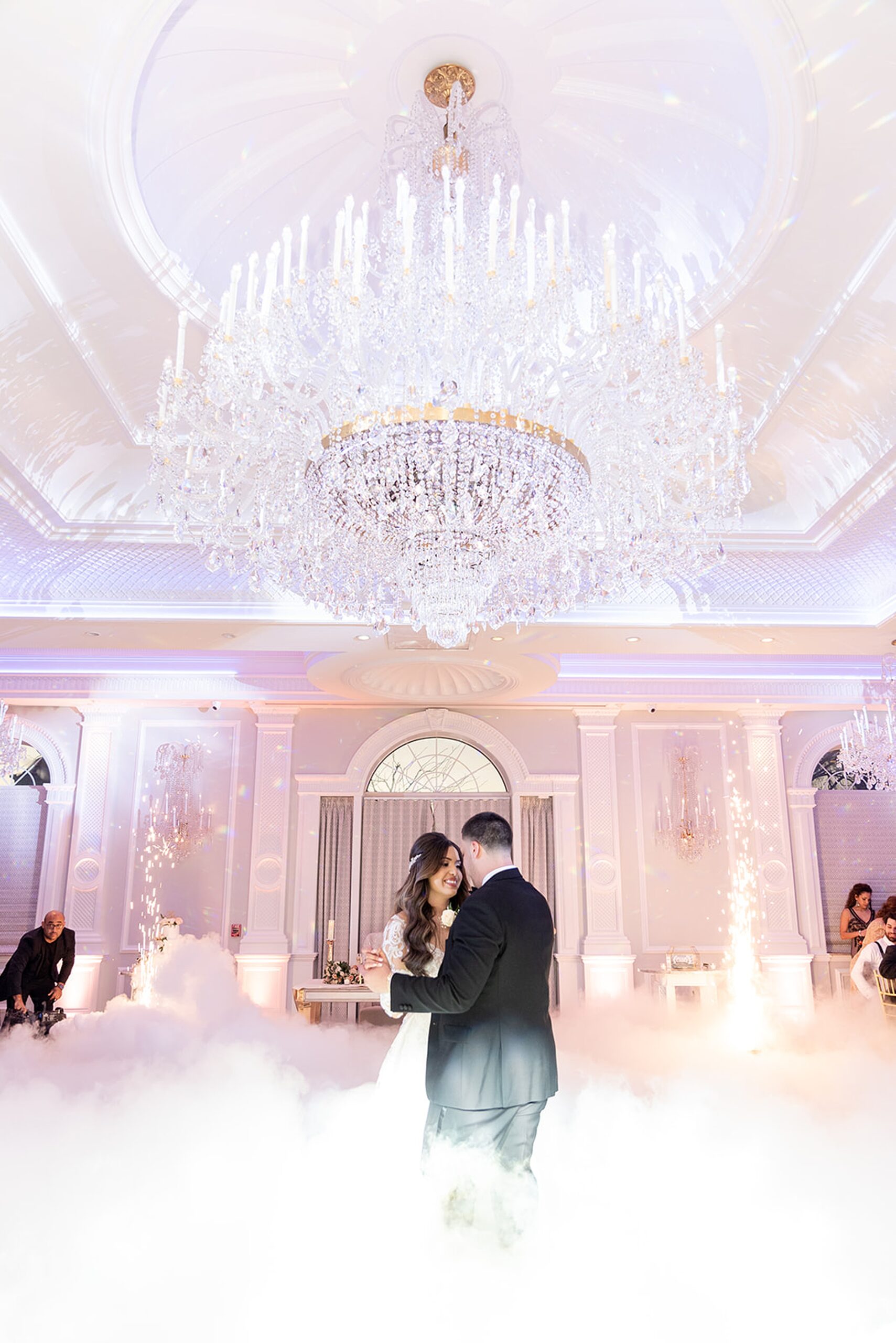 Newlyweds dance under a large crystal chandelier on a dance floor covered in fog with fireworks at the rockleigh wedding venue