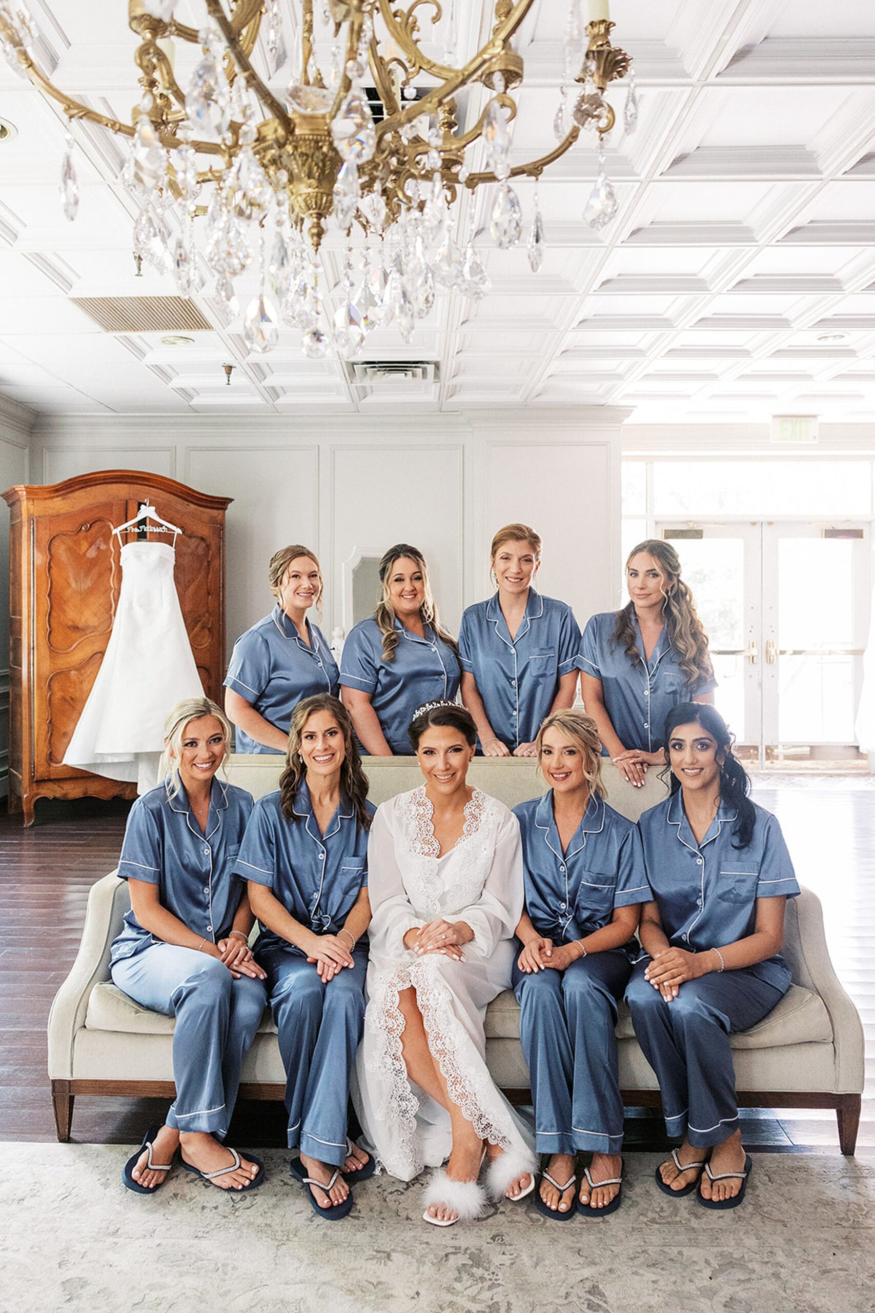 A bride sits in white pajamas on a couch surrounded by her bridesmaids in blue pajamas at a valley regency wedding