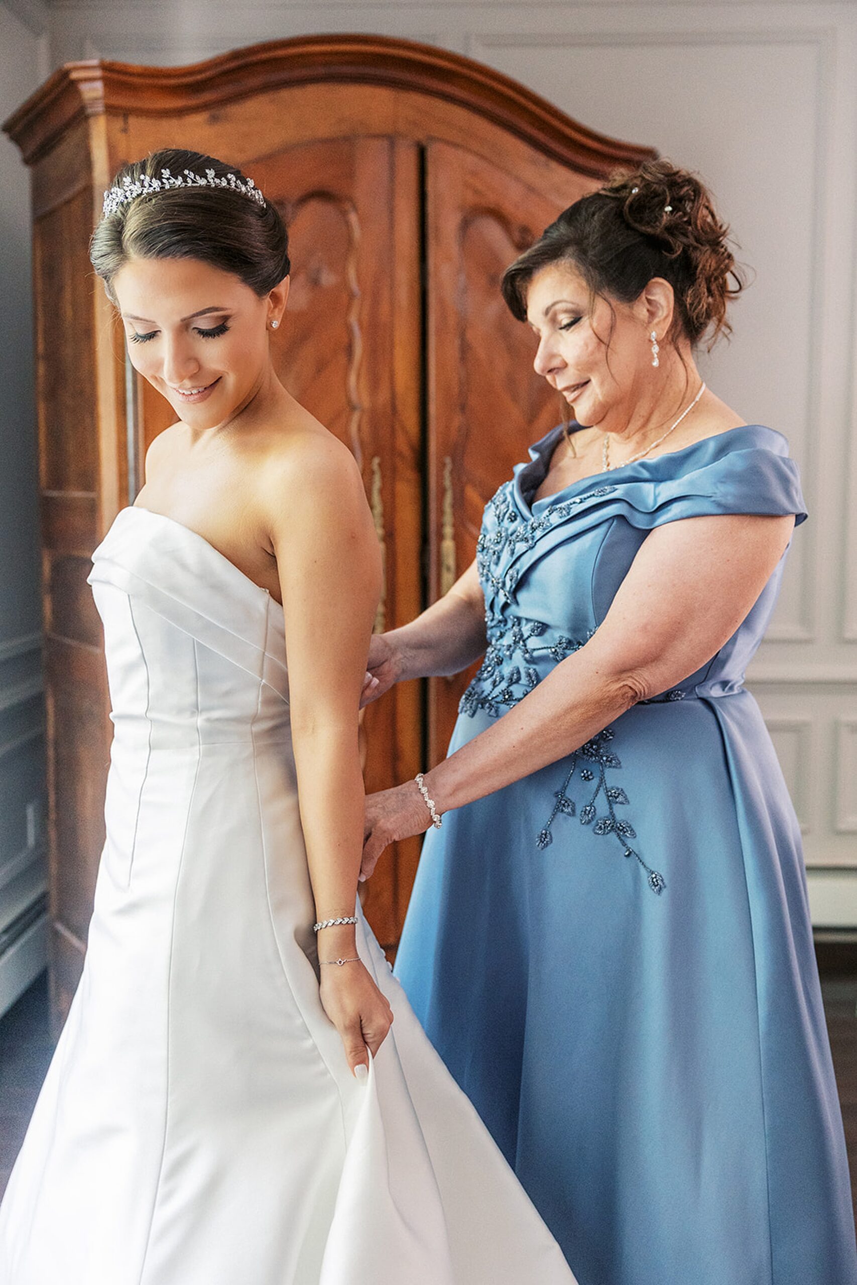 A mother in a blue dress helps zip up her daughter's silk wedding dress in front of a mirror