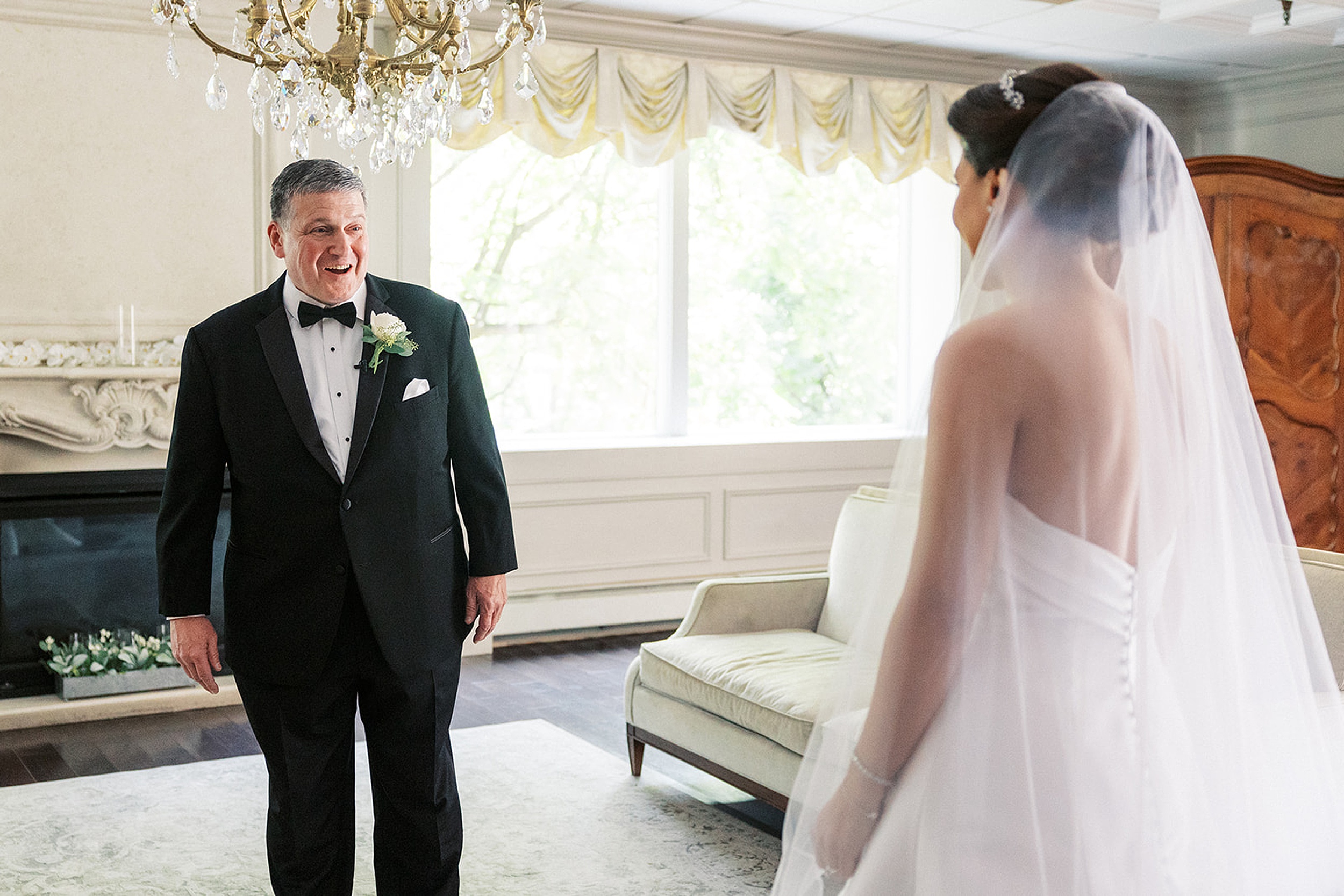 A father in a black tuxedo sees his daughter in her dress and veil for the first time under a crystal chandelier at a valley regency wedding