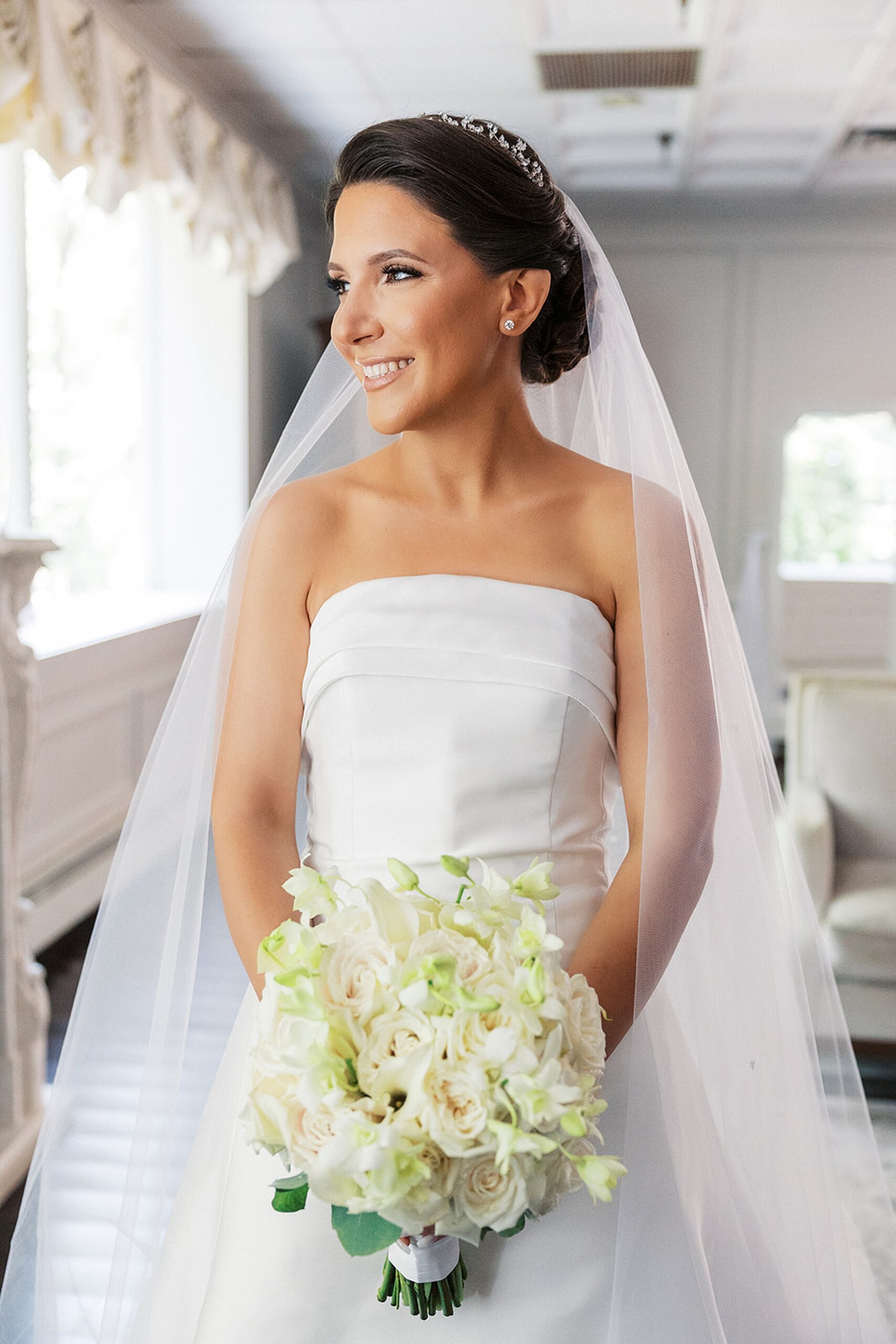 A bride smiles out a window while wearing her silk dress and holding her white rose bouquet at a valley regency wedding