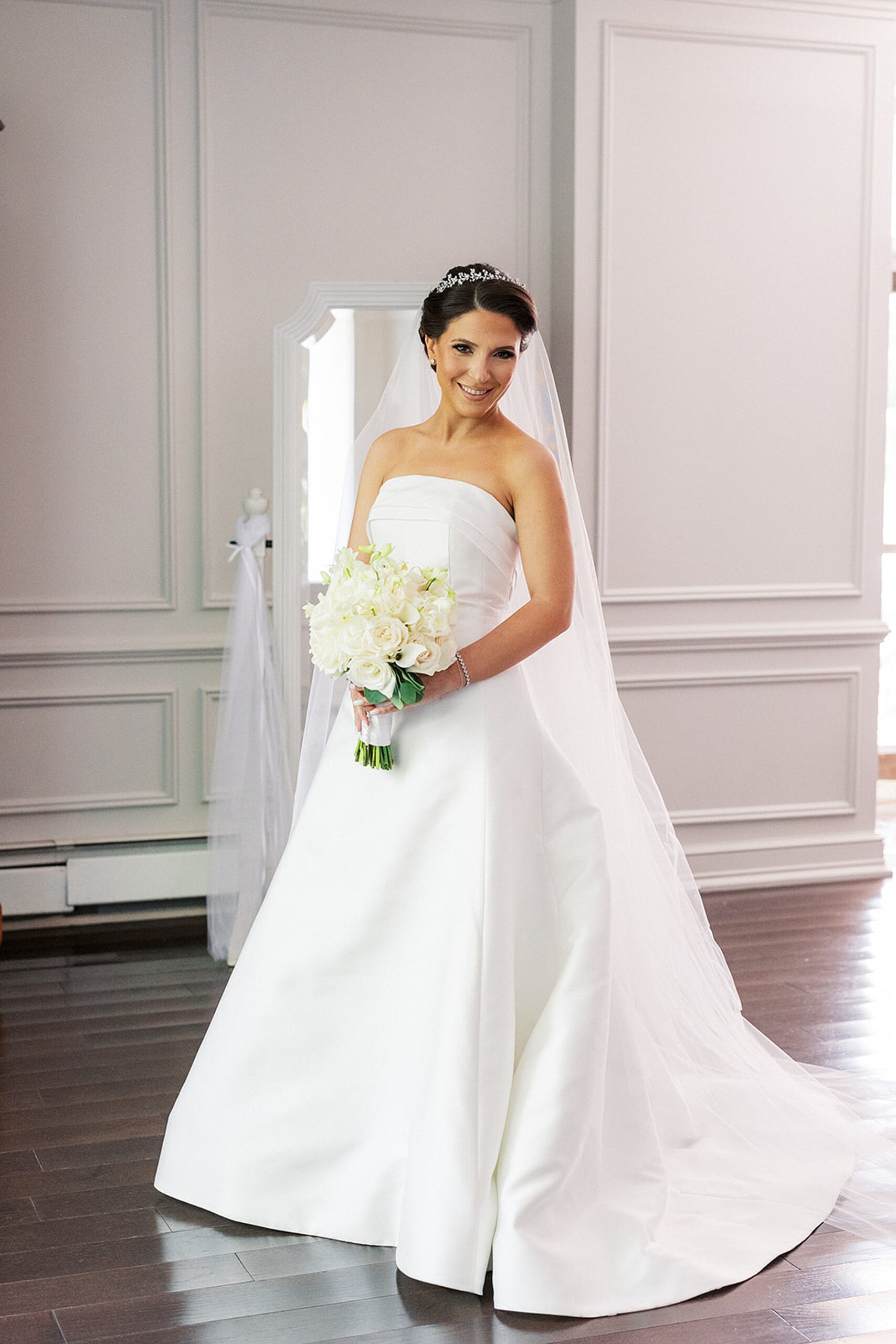 A bride stands in her dress in front of a mirror holding her white rose bouquet
