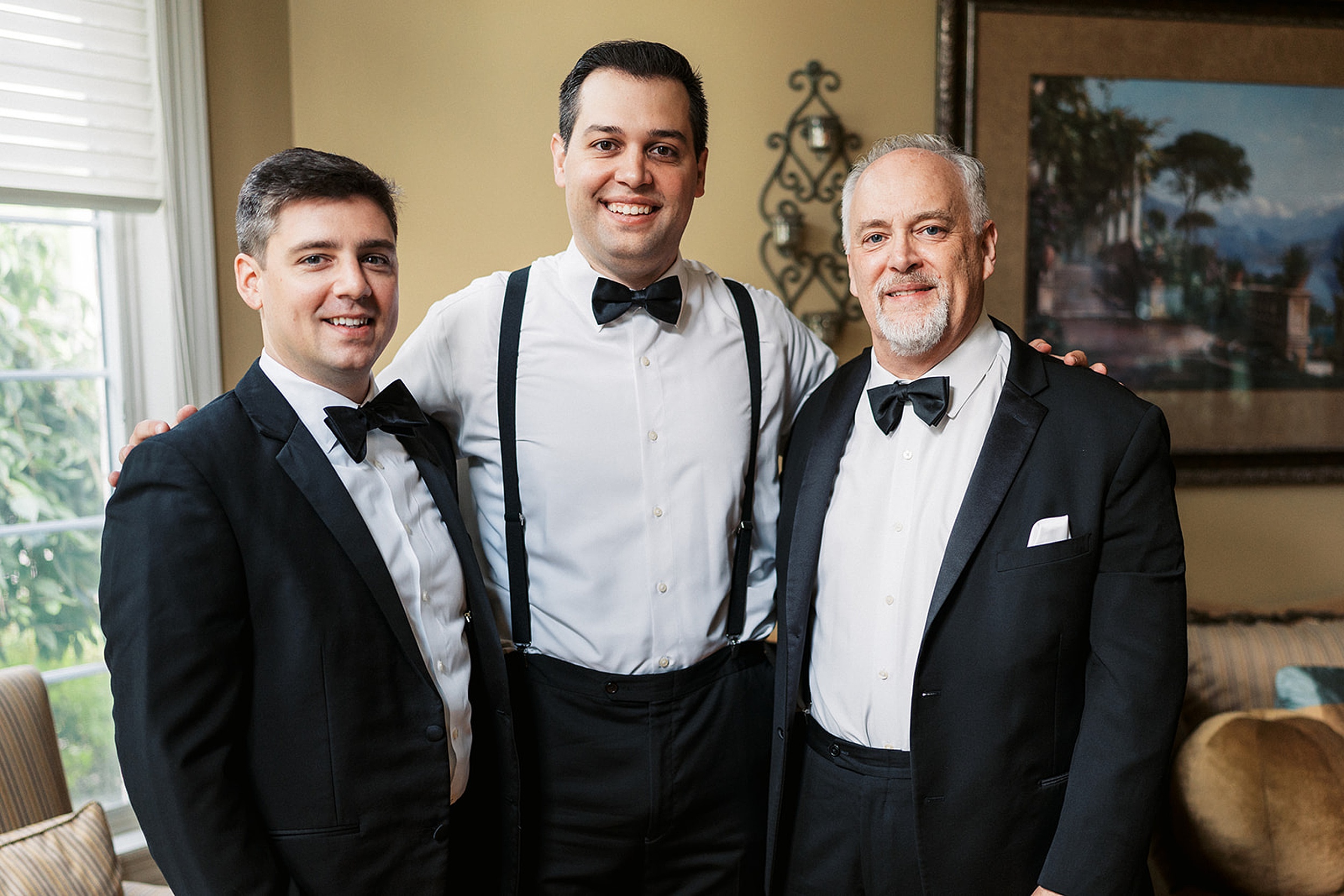 A groom stands in a room with his father and brother in black tuxedos and bowties by a window