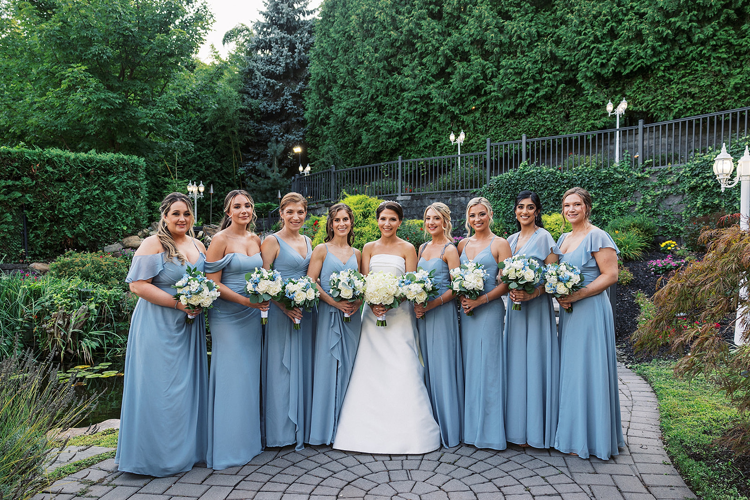 A bride stands in a garden by a pond with her bridal party wearing blue dresses and holding white rose bouquets at a valley regency wedding