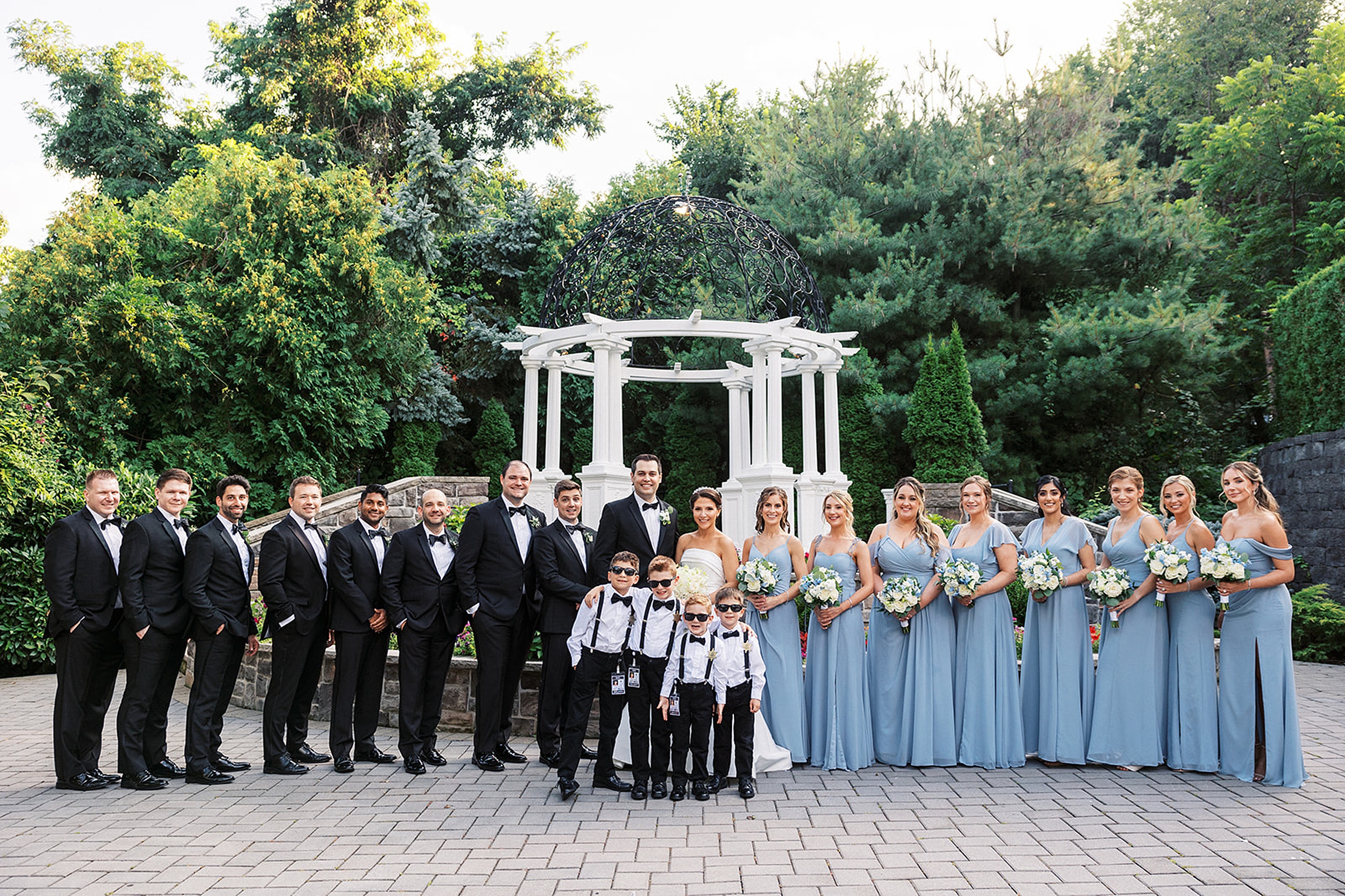 Newlyweds stand together with their bridal party and security kids in a garden by a large white gazebo