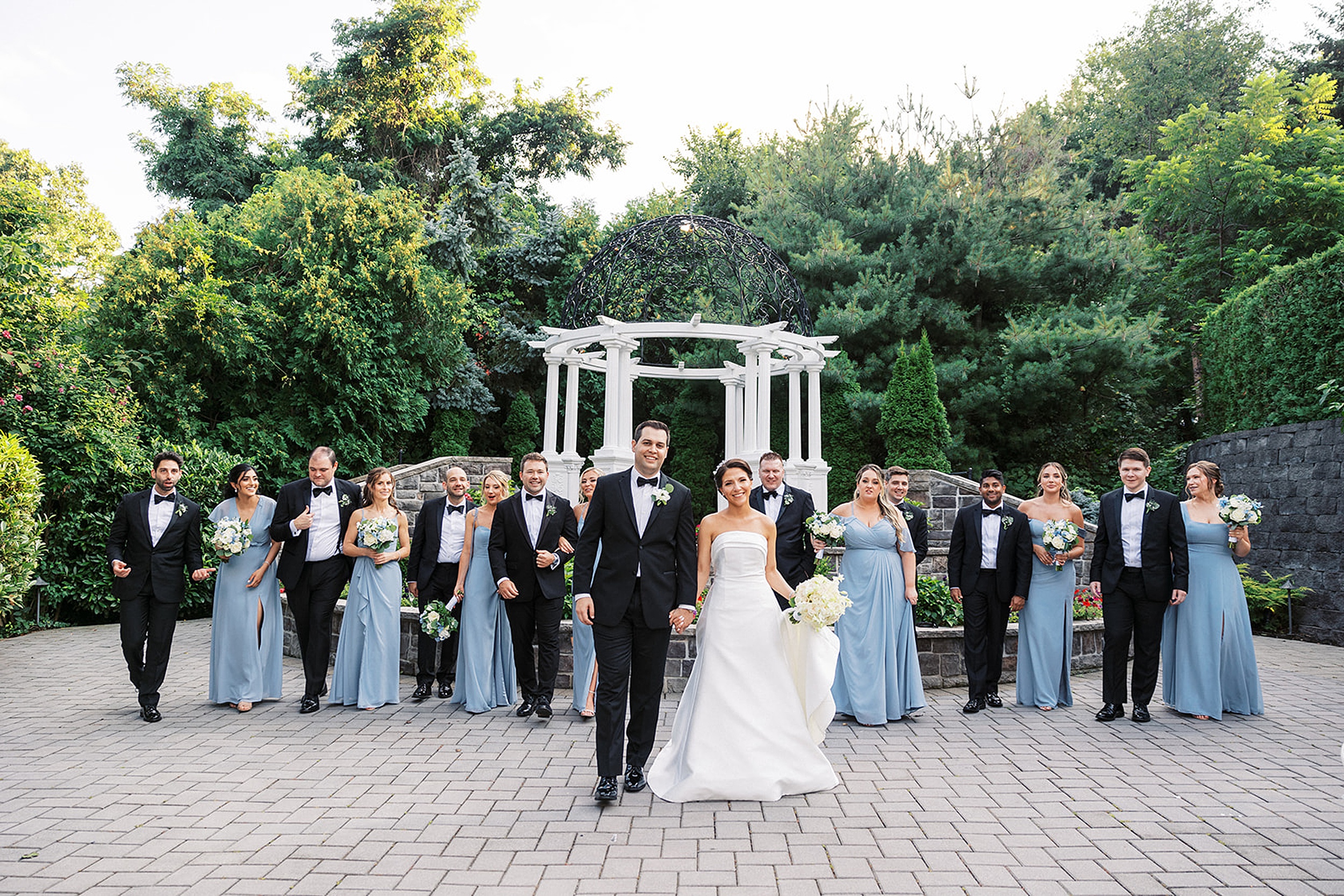 Newlyweds hold hands while walking through a garden surrounded by their large wedding party in black suits and blue dresses