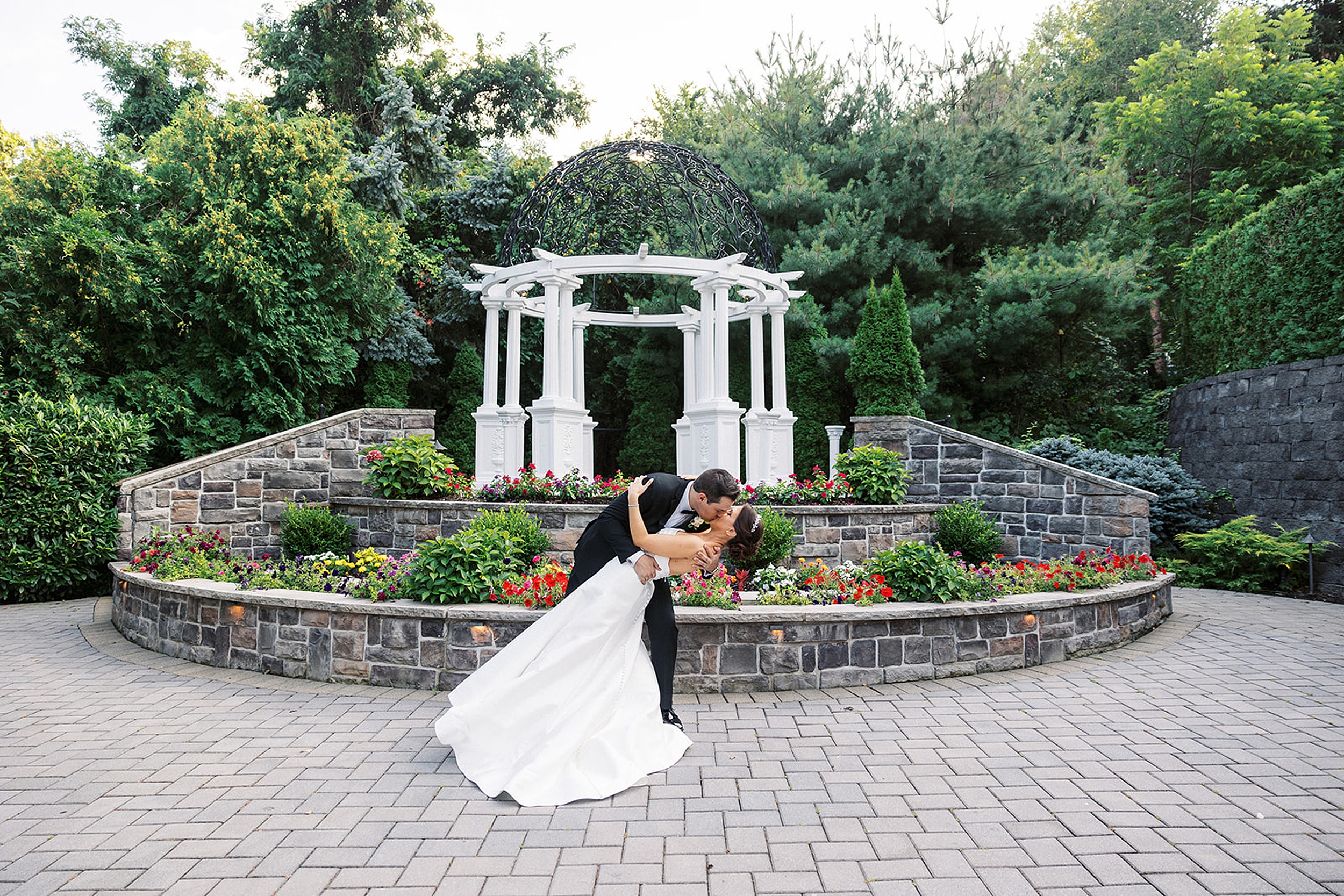 A groom dips and kisses his bride in front of a large raised garden bed with a white gazebo above it at a valley regency wedding