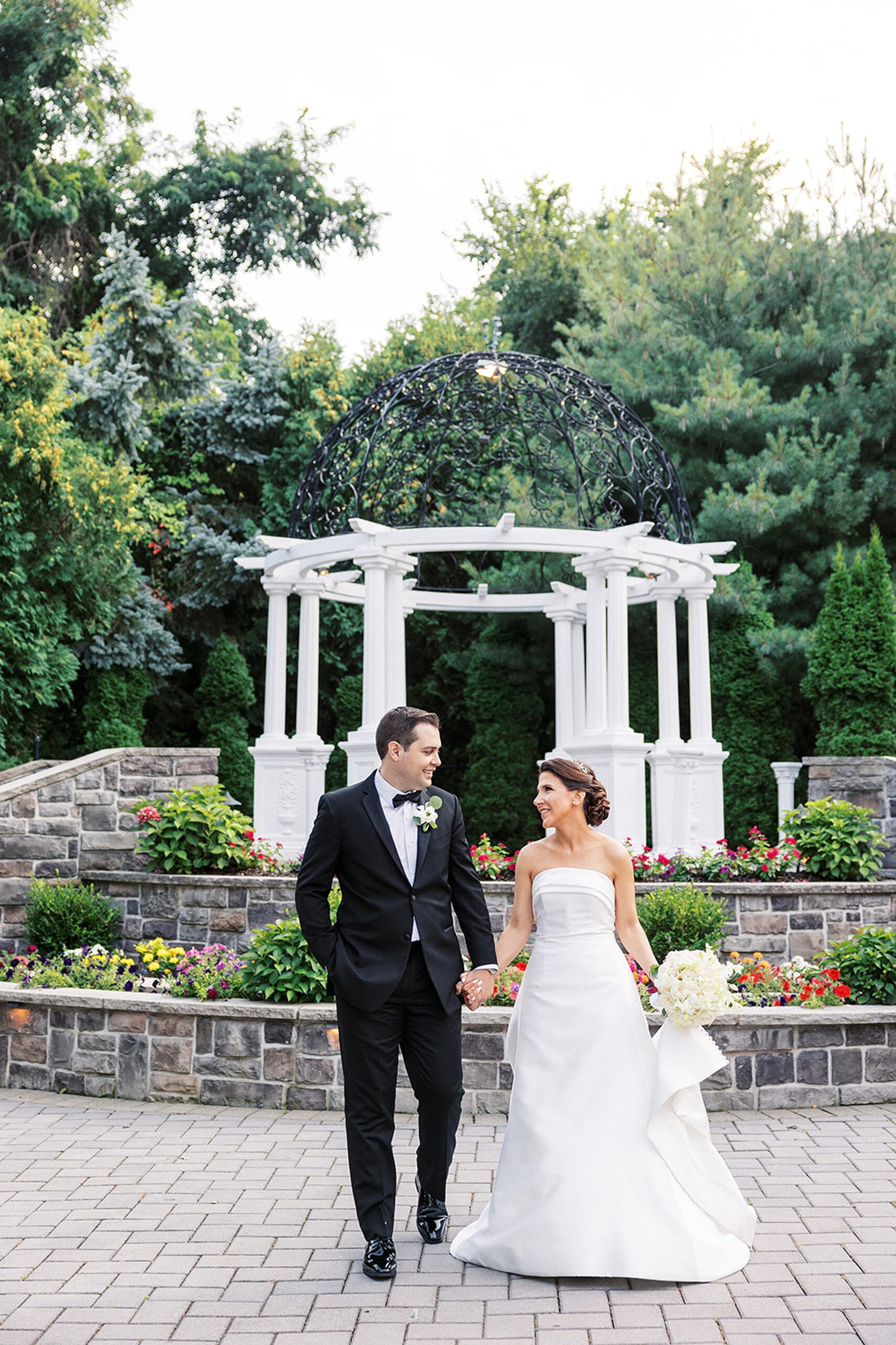 Newlyweds hold hands while walking through a garden with a large white gazebo