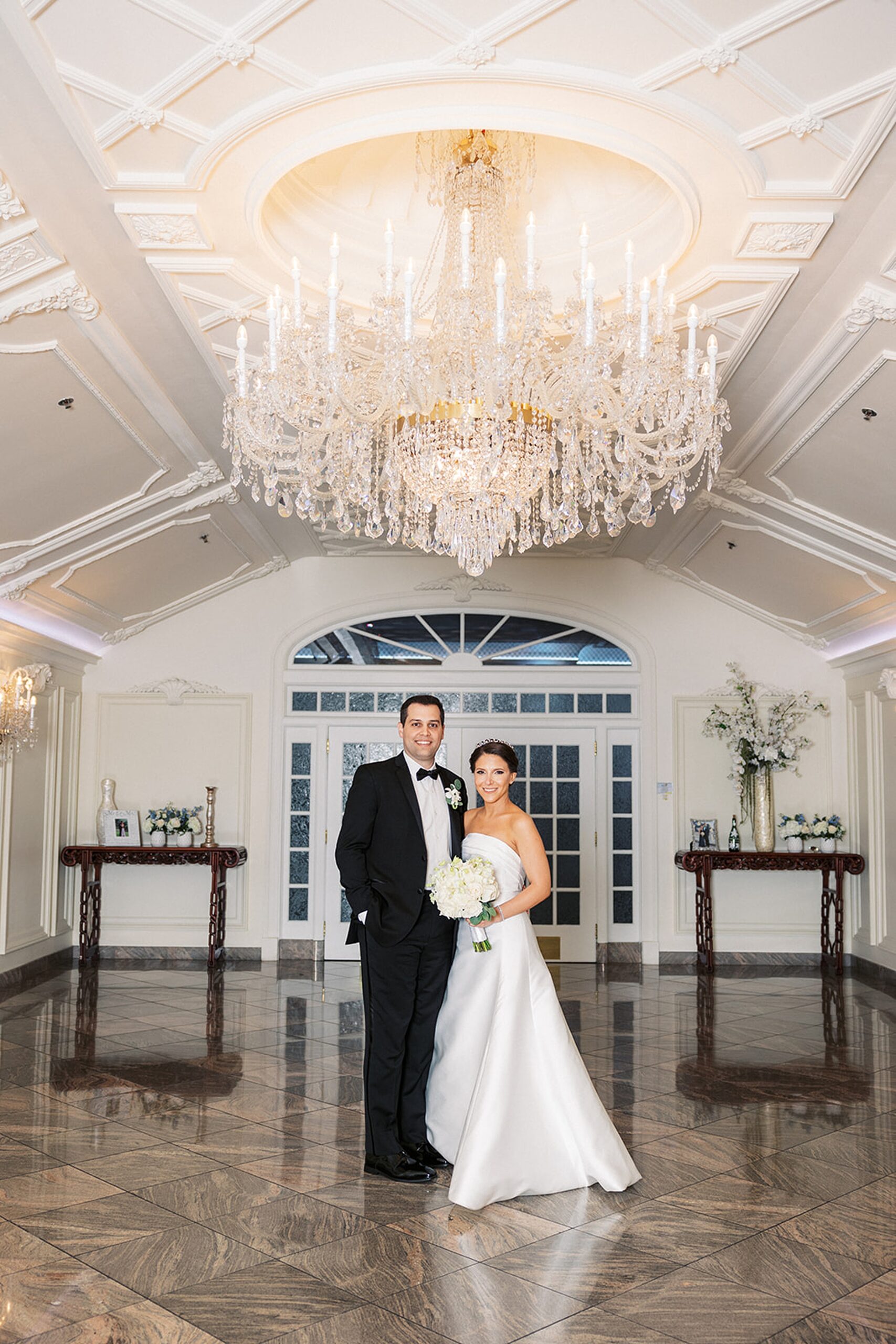 Newlyweds stand together under a large crystal chandelier at the entrance to their wedding reception