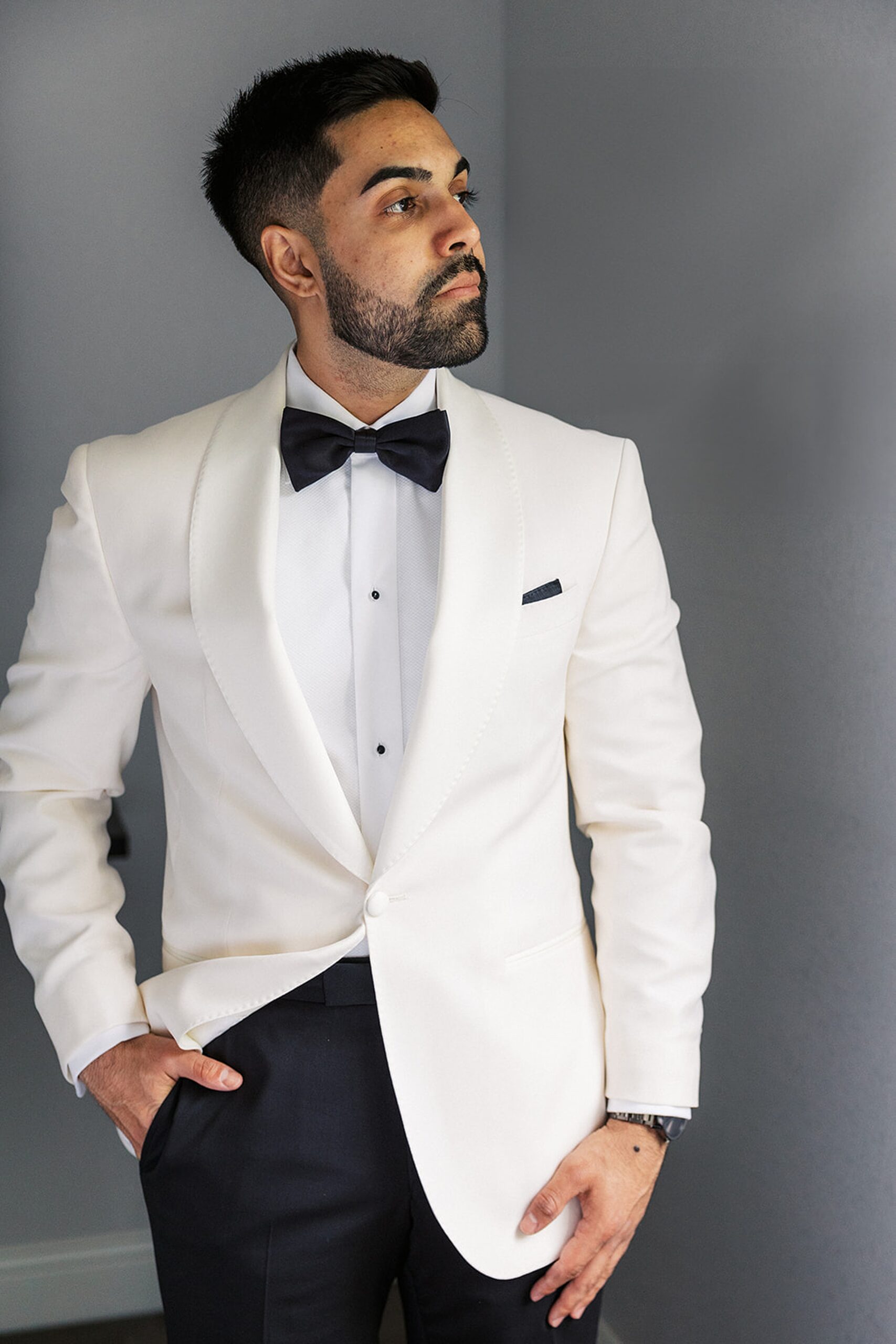 A groom in a white tuxedo jacket and black pants gazes out a window