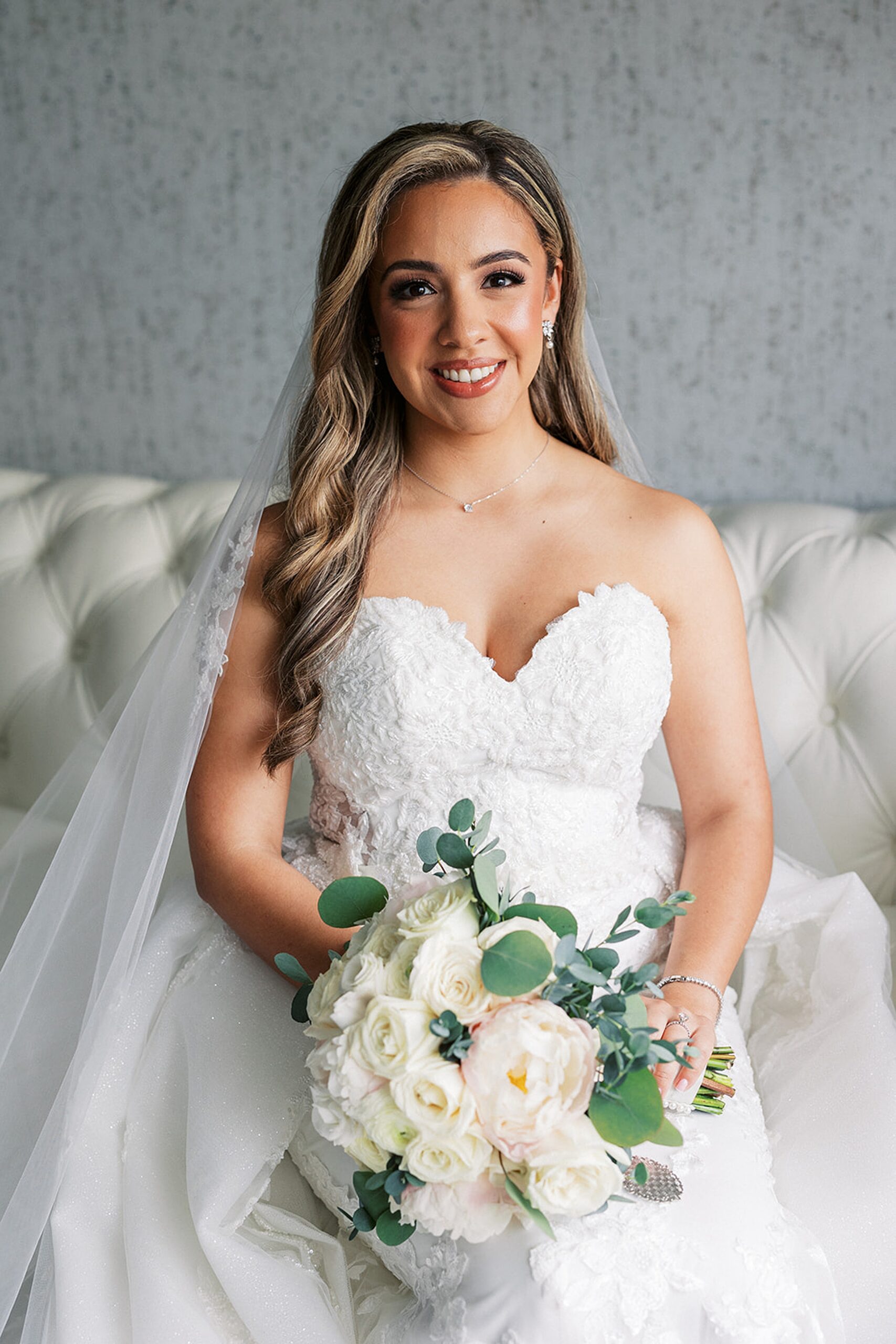 A bride sits on a couch in a white lace embroidered dress holding her white rose bouquet