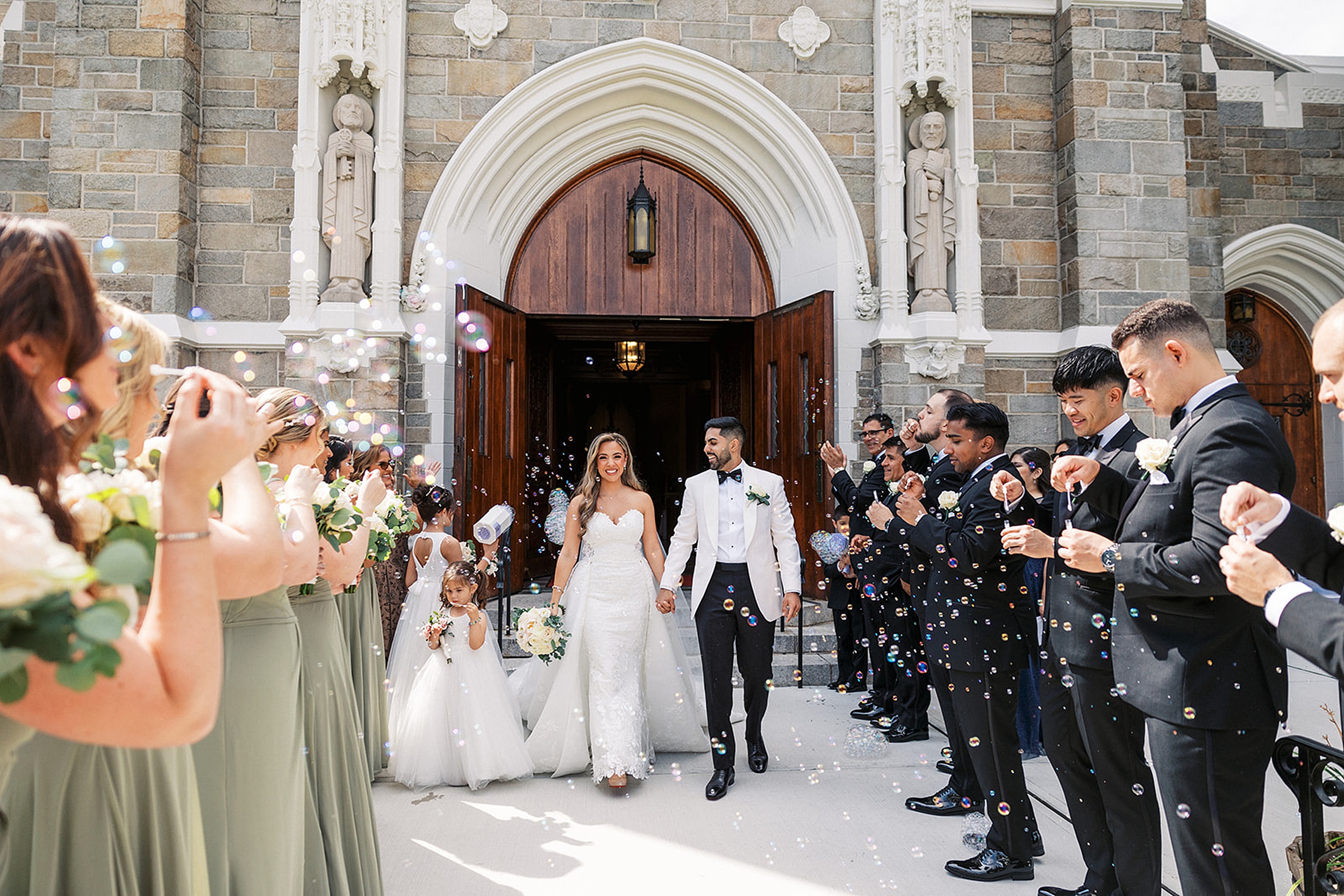 Newlyweds leave their wedding ceremony through large wooden doors while their wedding party blows bubbles over them at a valley regency wedding