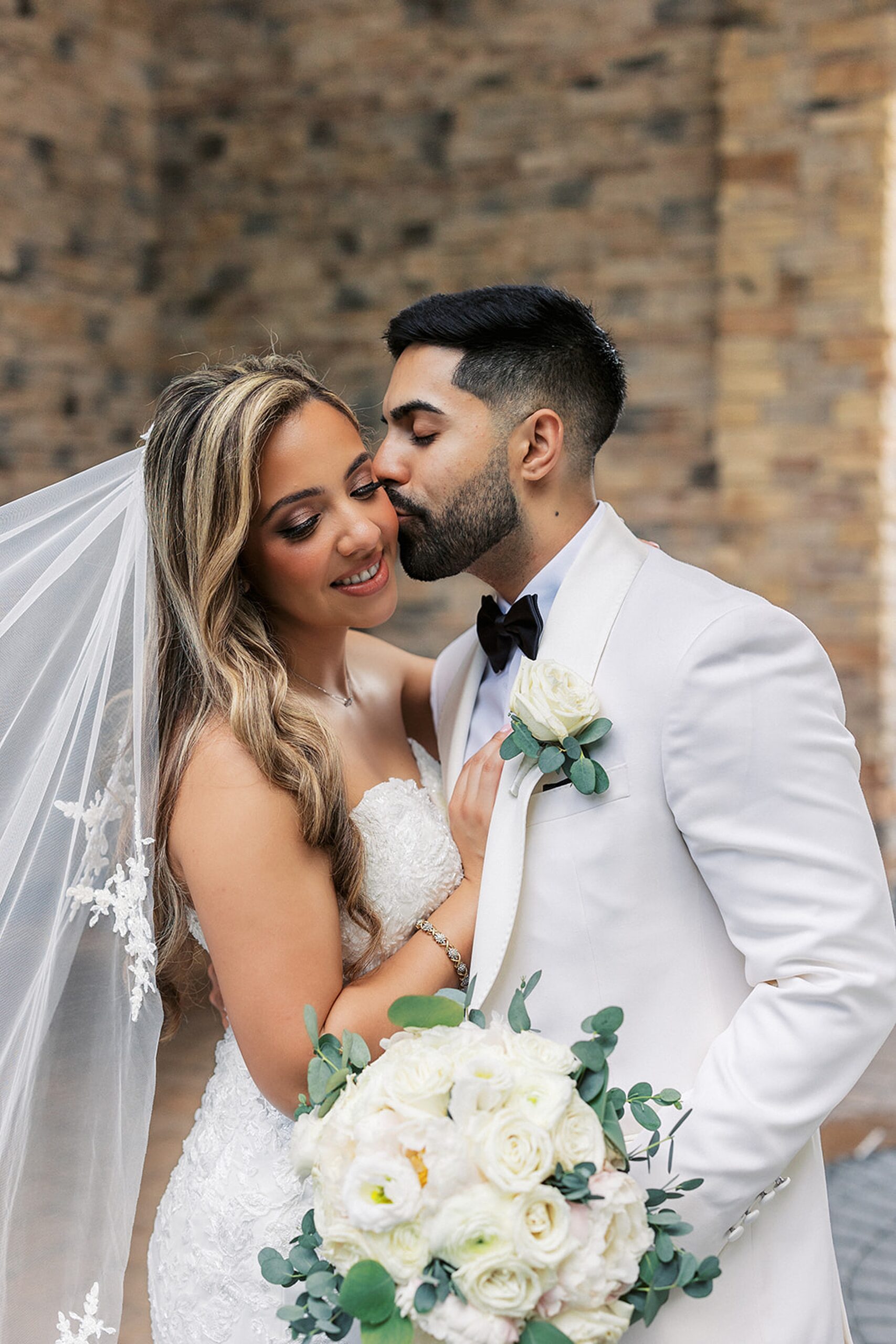 A groom kisses his bride while wearing a white tuxedo jacket and holding her white rose bouquet as her veil flows in the wind at a valley regency wedding
