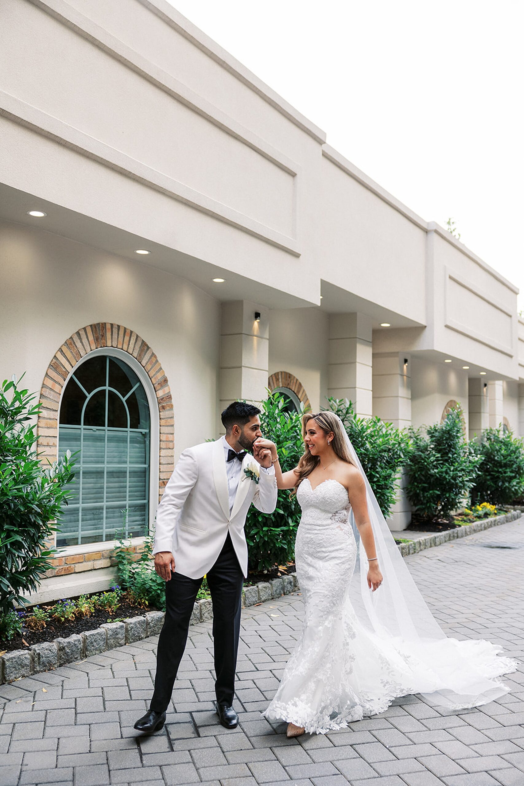 A groom in a white tuxedo and black pants kisses the hand of his bride as they walk through a garden behind the valley regency wedding venue