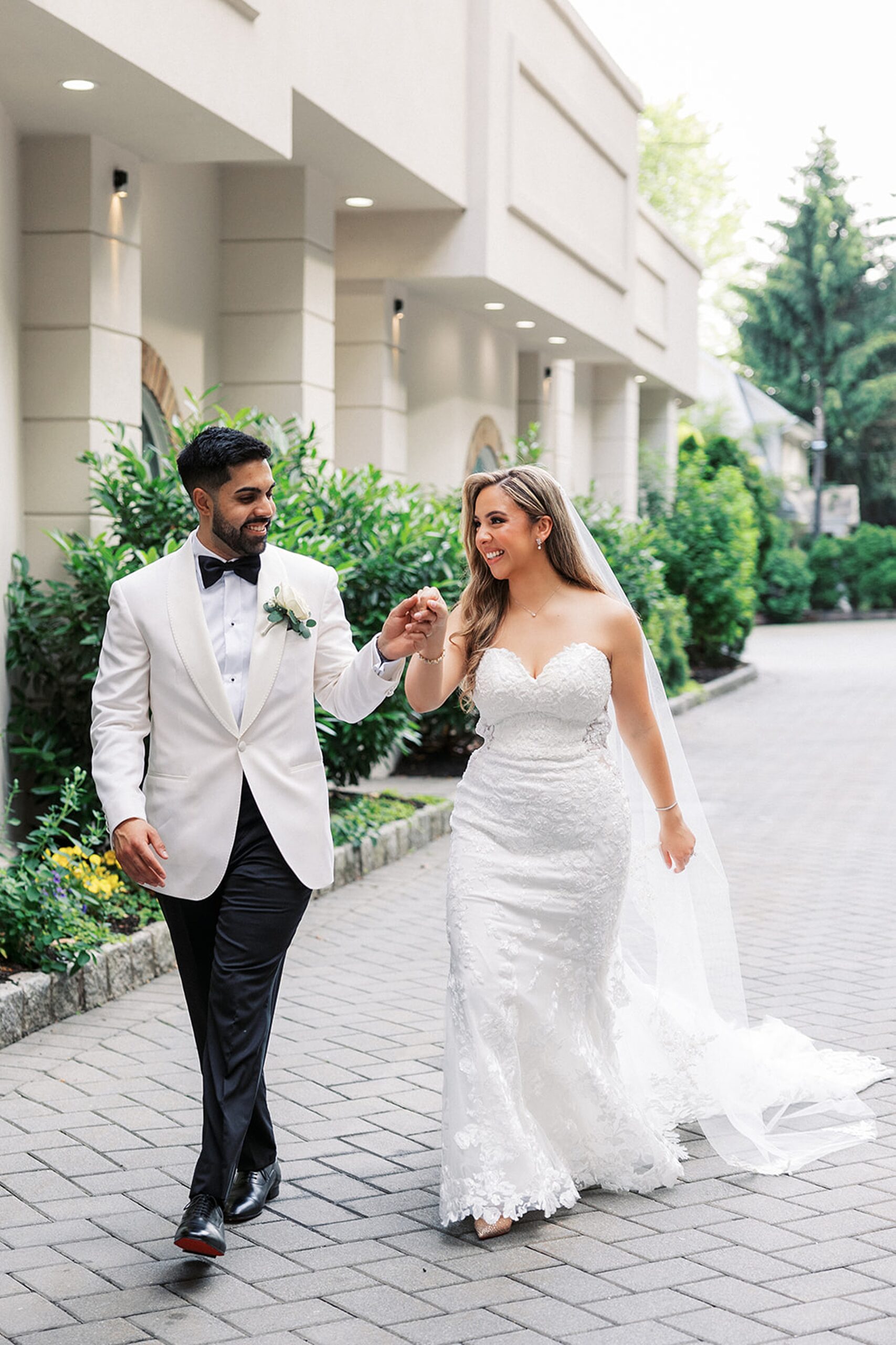 A groom in a white tuxedo walks his bride through a garden on a stone path while holding her hand