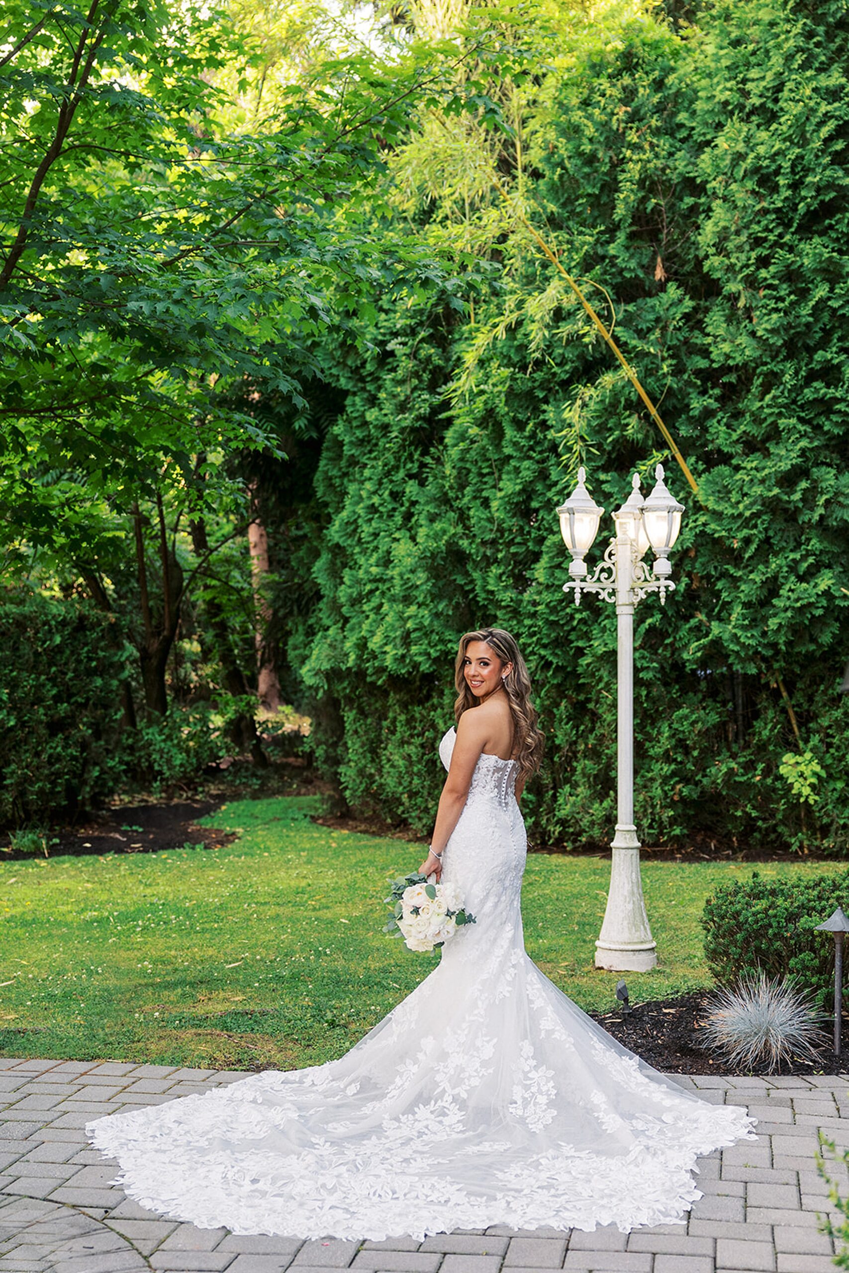 A bride with a long lace embroidered train stands in a garden with a white lamp post at a valley regency wedding