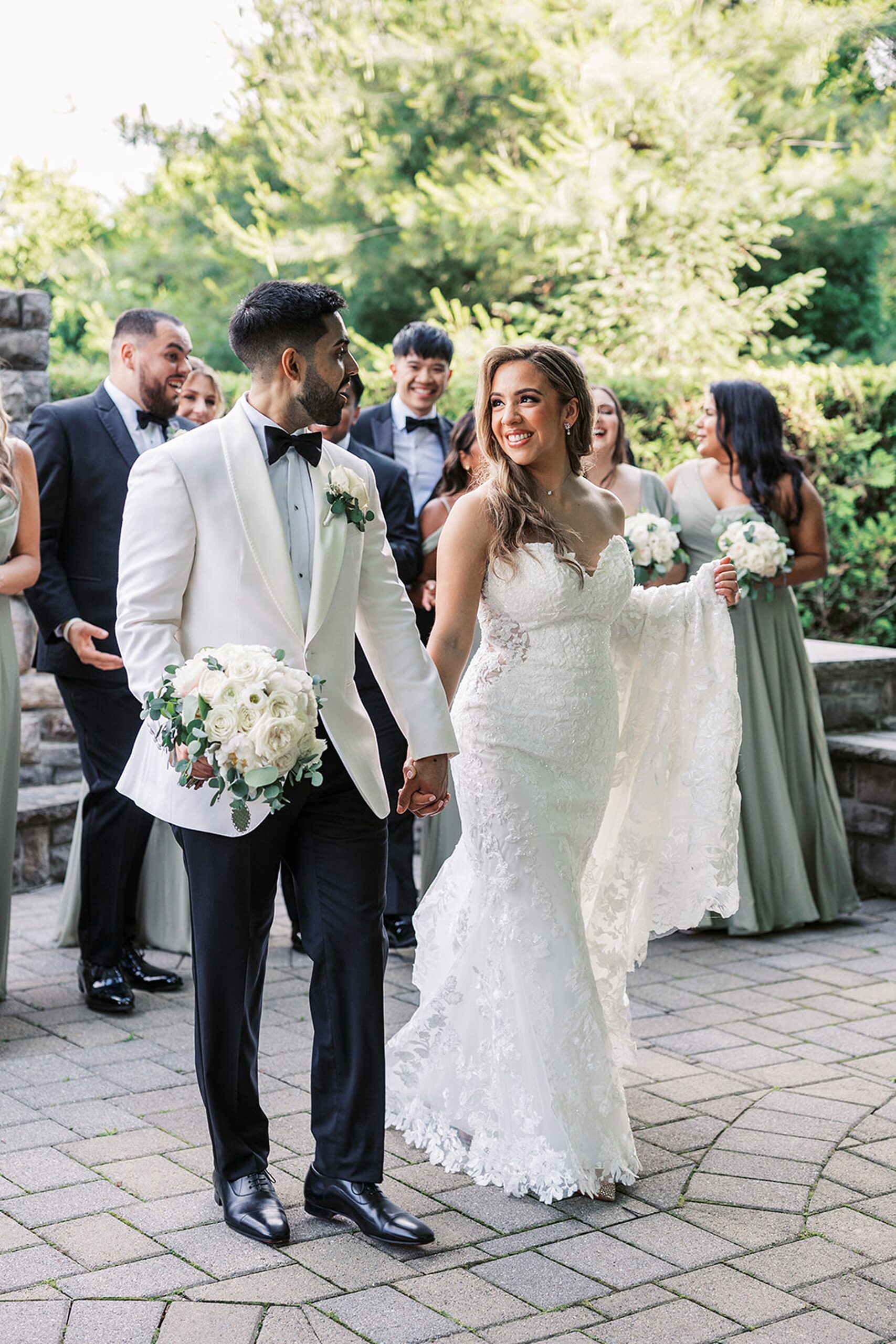 Newlyweds hold hands while walking through a garden as their wedding party laughs and celebrates behind them