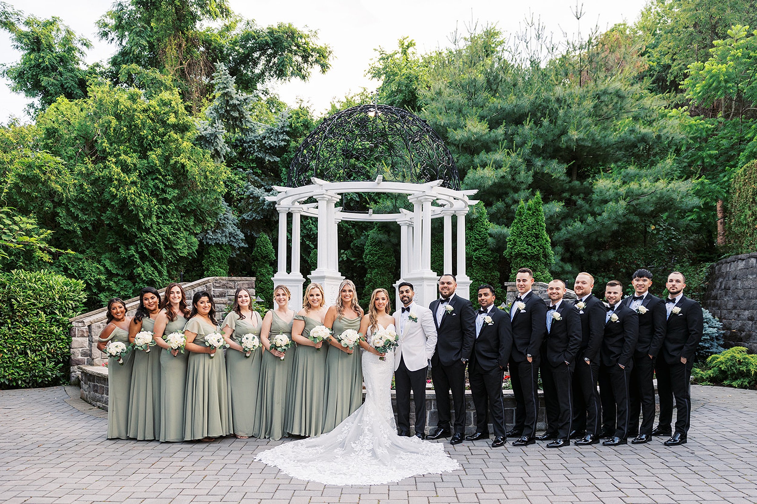 Newlyweds stand in a garden by a gazebo while surrounded by their wedding party