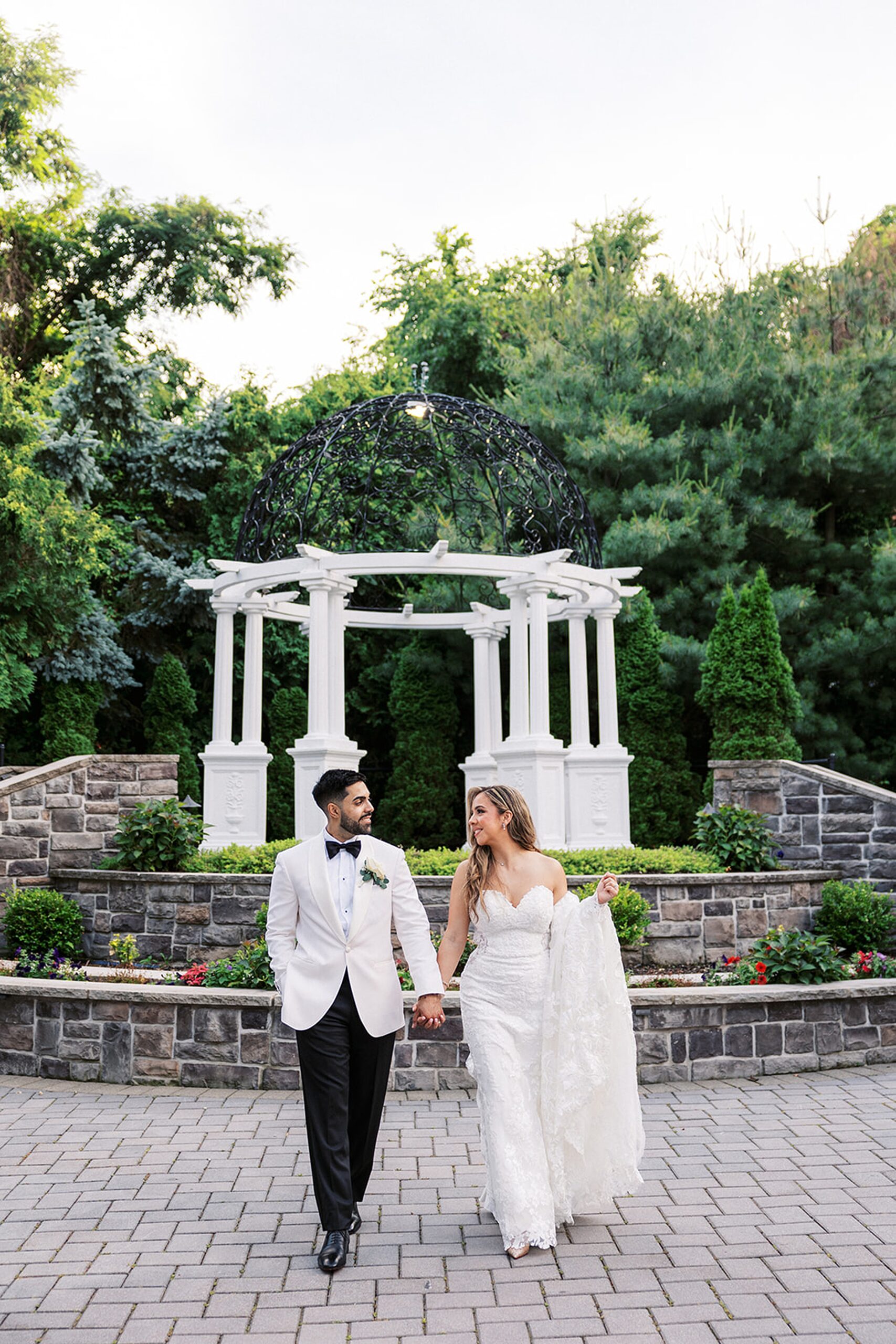 Newlyweds hold hands while walking through a garden path with a large white gazebo