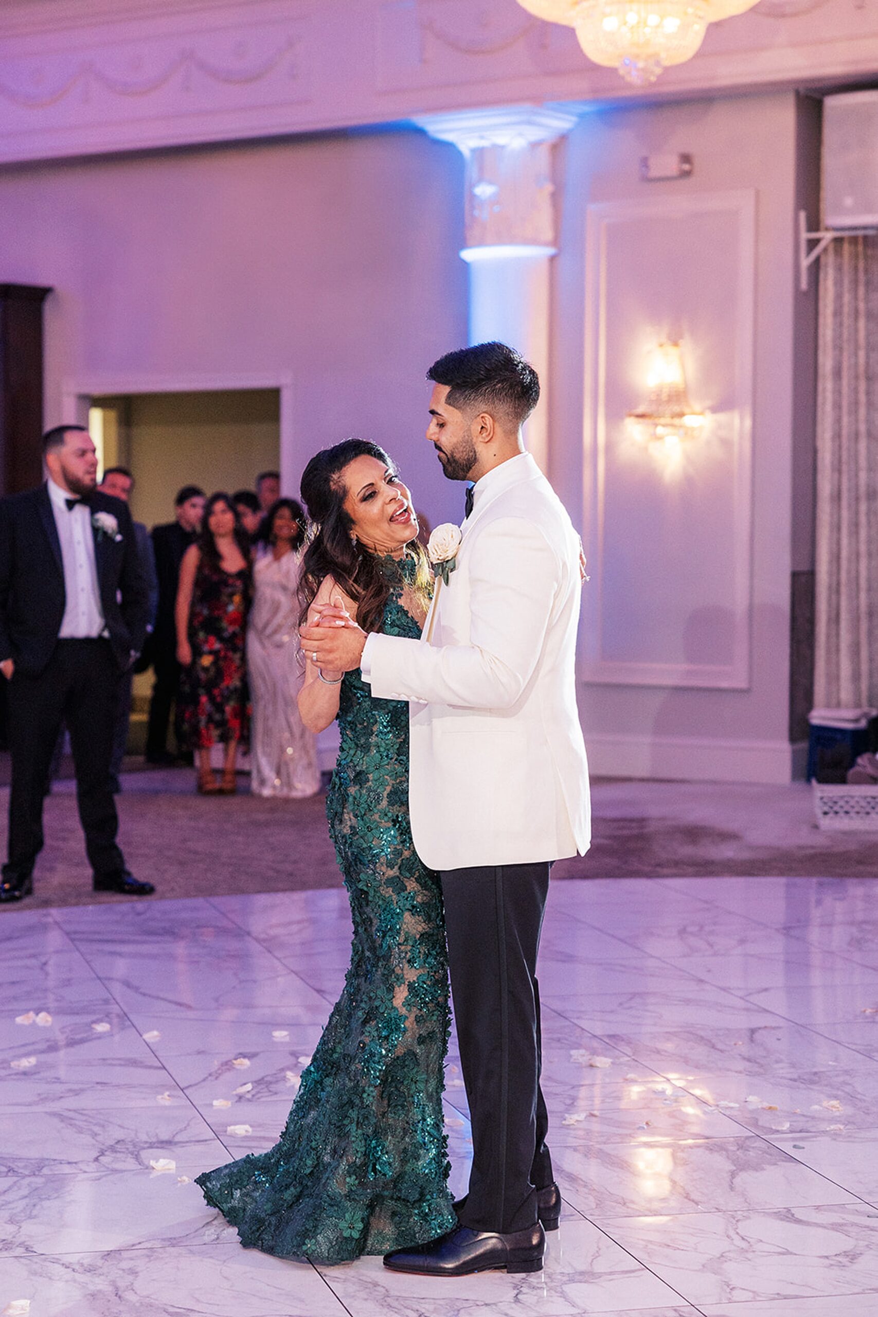 A groom in a white jacket dances with his mother in a green lace dress on a marble dance floor at a valley regency wedding