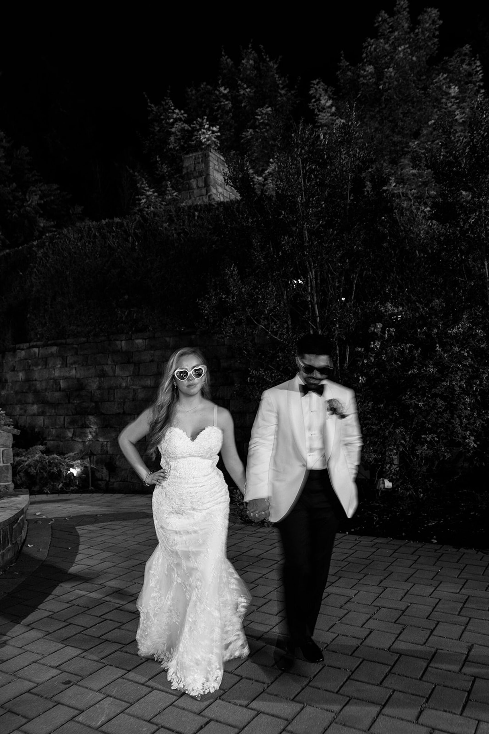 Newlyweds wear large sunglasses while walking hand in hand through a garden patio