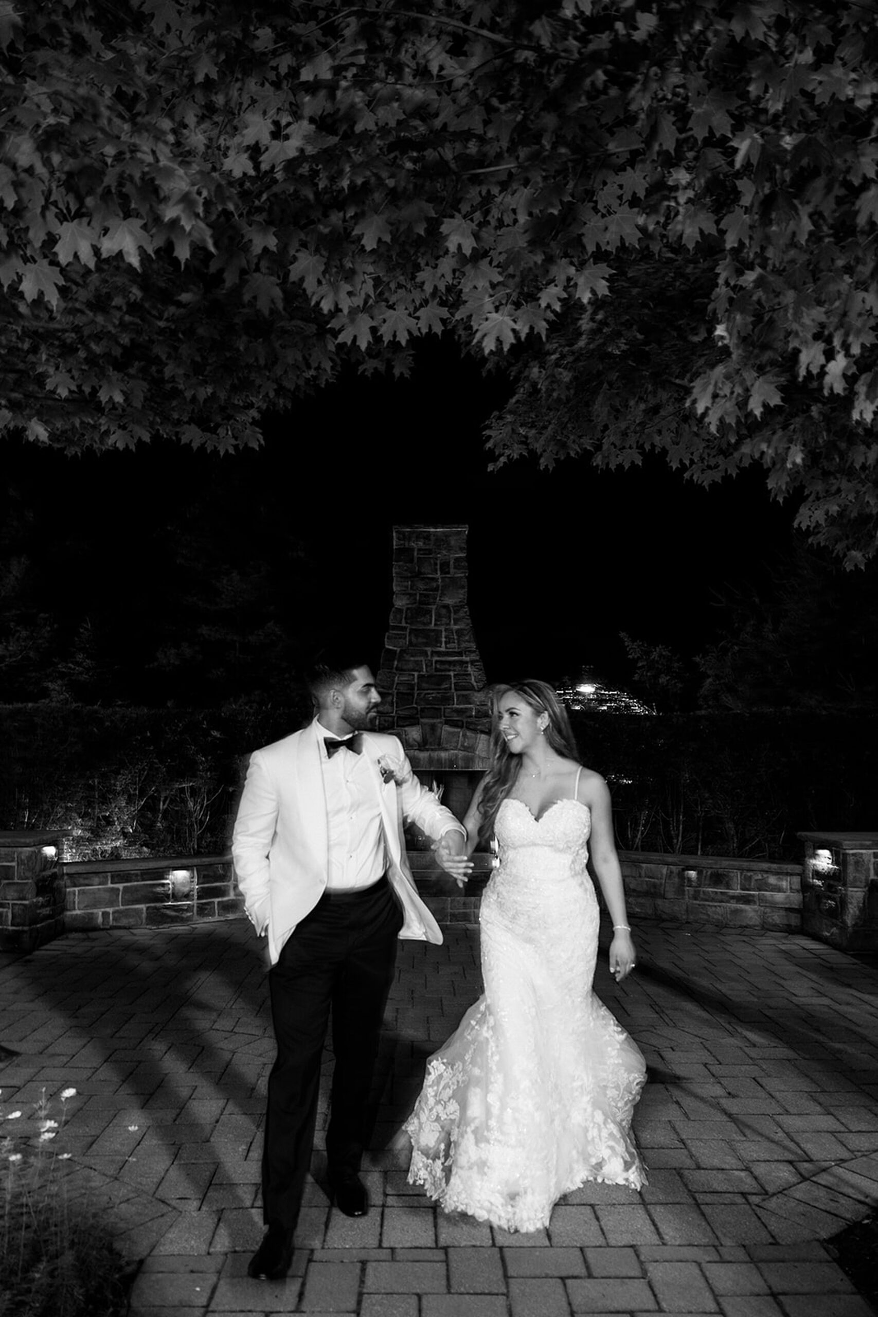 Newlyweds walk hand in hand through a paved patio garden with a stone fireplace at their valley regency wedding