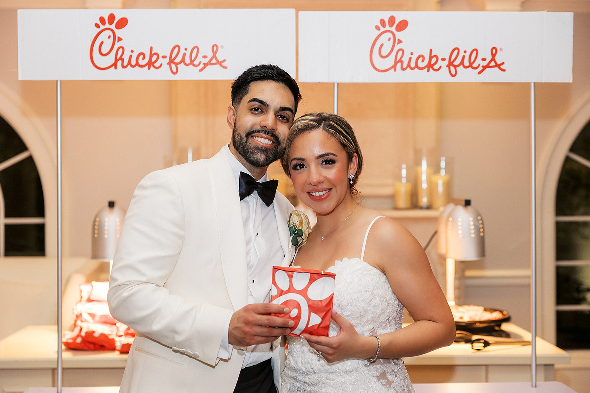 Newlyweds stand holding a chick-fil-a sandwhich in front of their chick-fil-a station at their valley regency wedding
