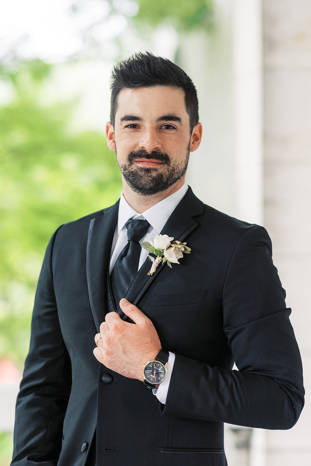 A groom in a black tuxedo holds his lapel while standing outside