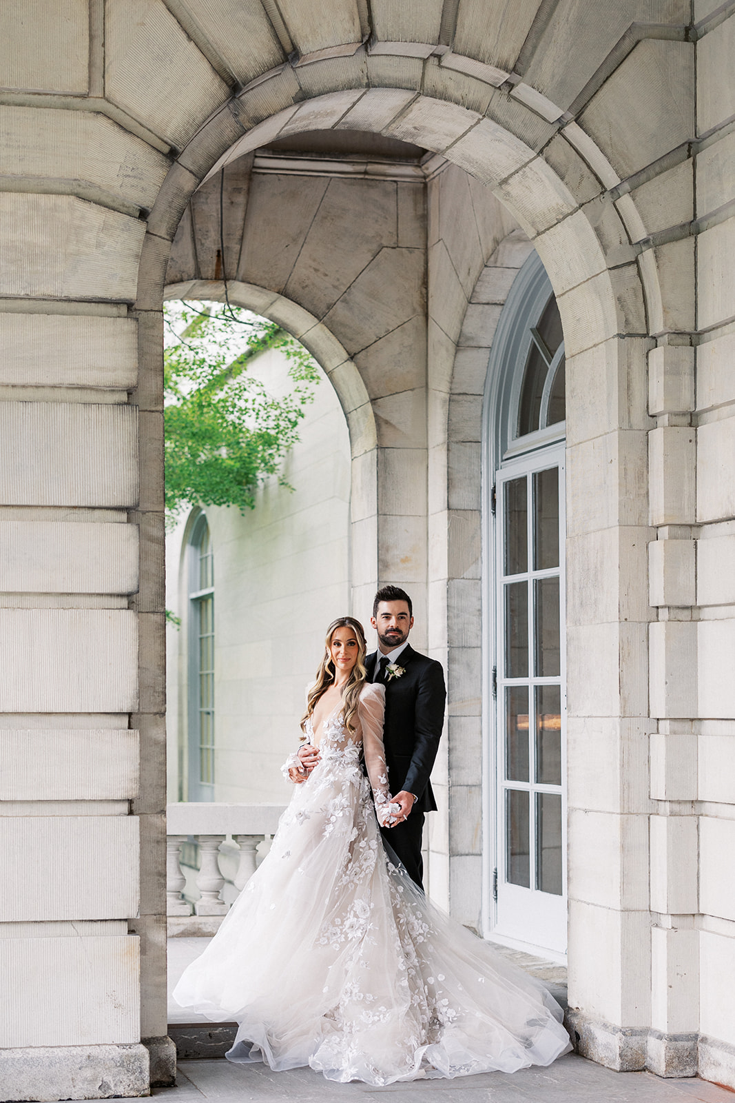 Newlyweds stand together holding hands under a large arch on a terrace