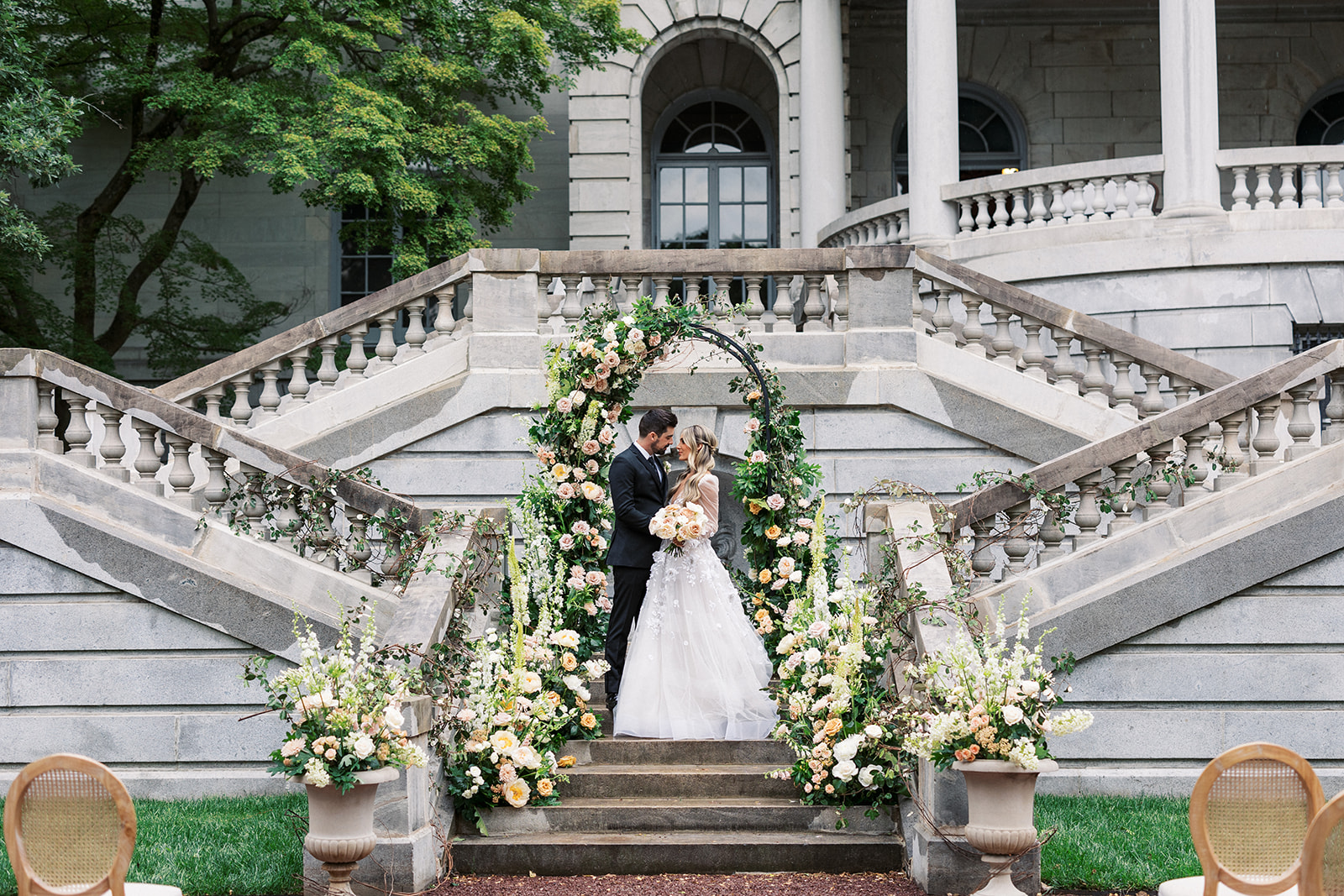 Newlyweds stand under a large flower arch on the ornate stairs of the Elkins Estate