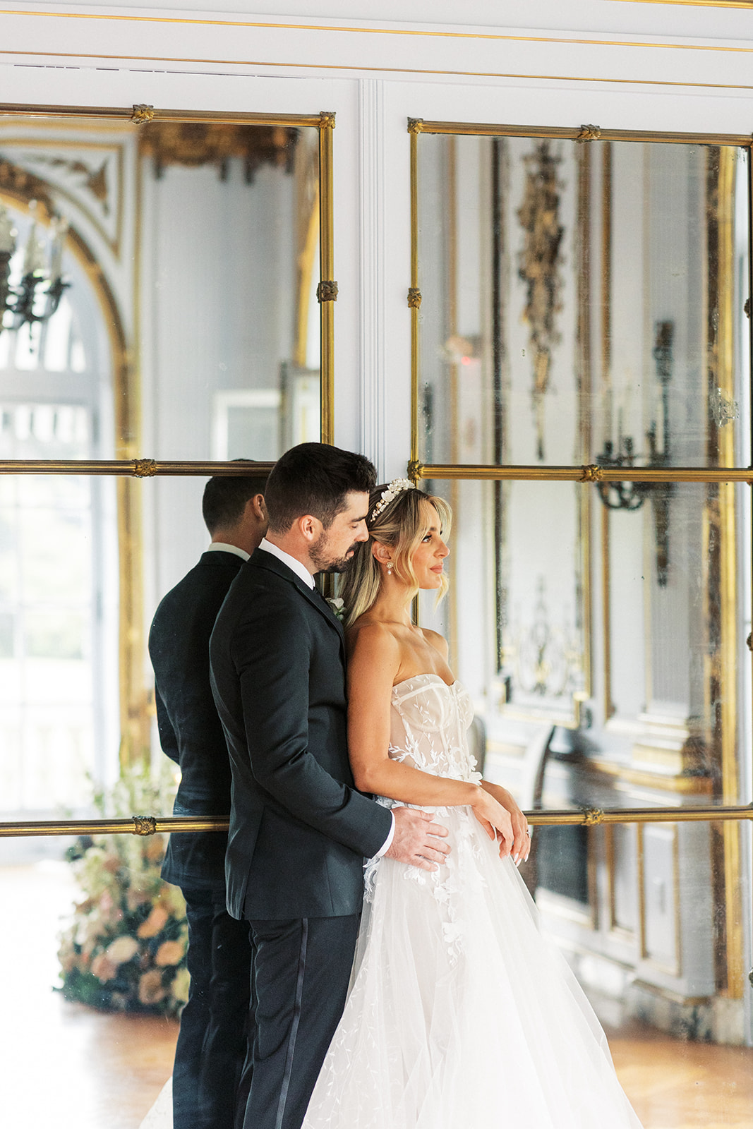 A bride leans into her groom as they stand in front of a large mirror with gold frame