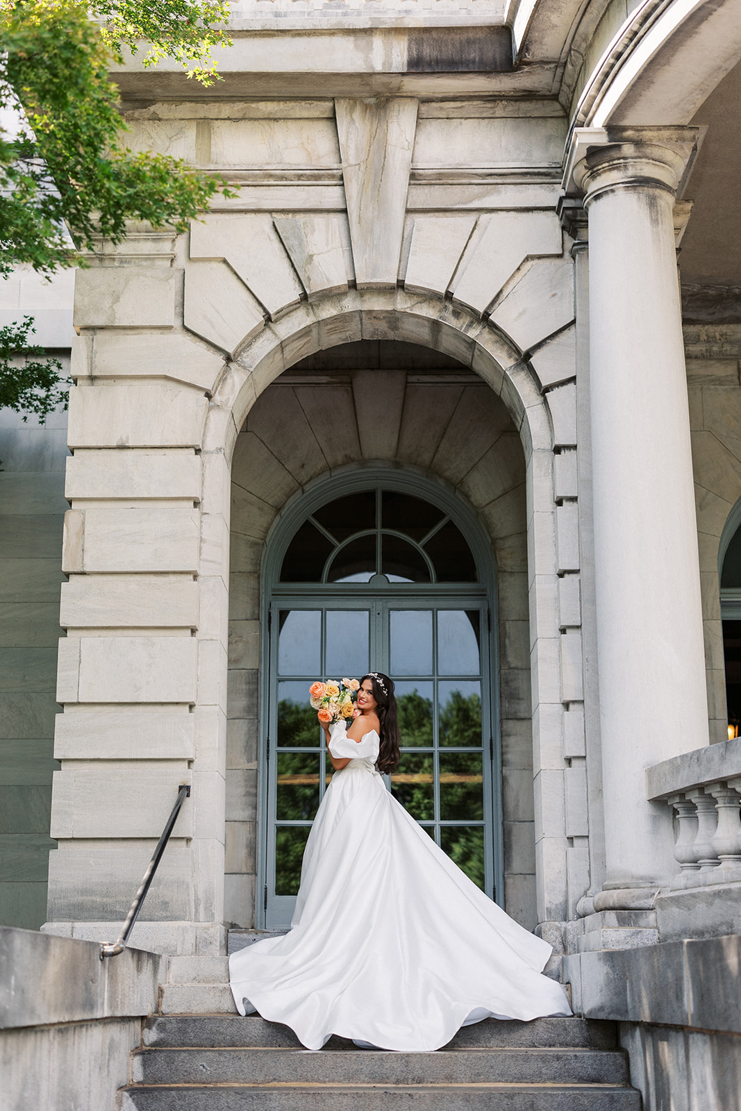 A bride holds her large bouquet up to her cheek while standing in a large arch at the top of an outdoor set of stairs