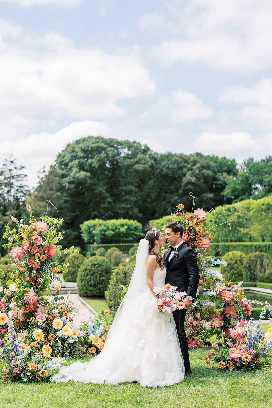 Newlyweds lean in for a kiss while standing in a colorful garden at the Oheka Castle