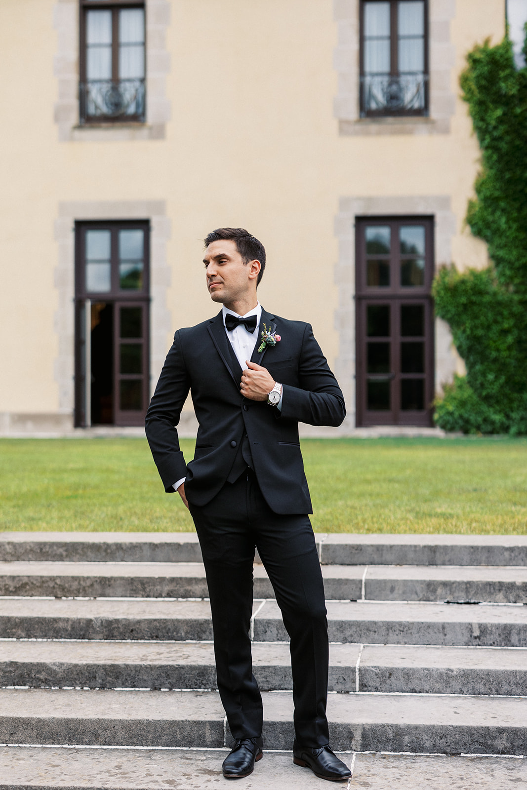 A groom in a black tuxedo holds his lapel while standing on garden steps