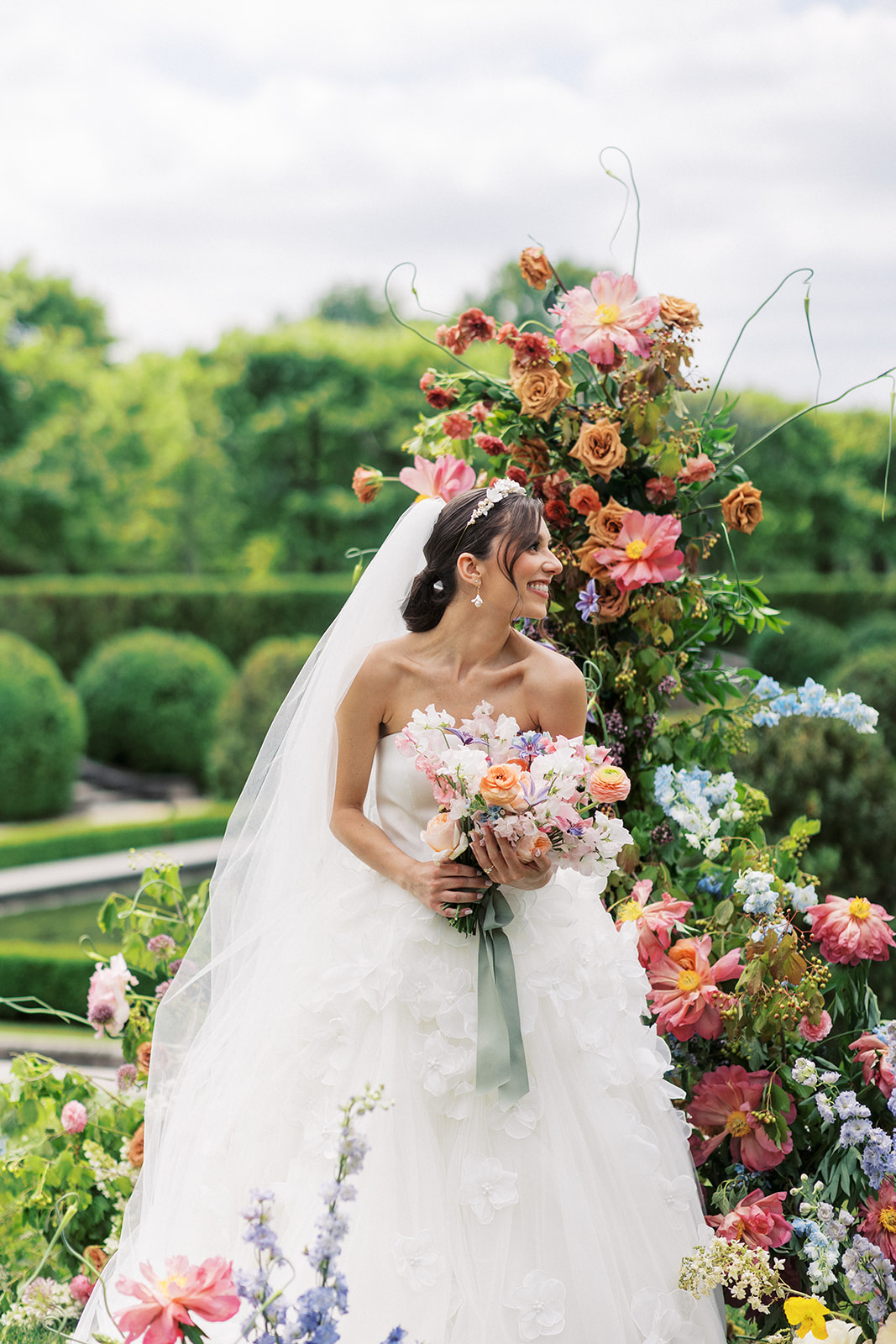 A bride smiles over her shoulder while standing amongst a large flower display in a garden