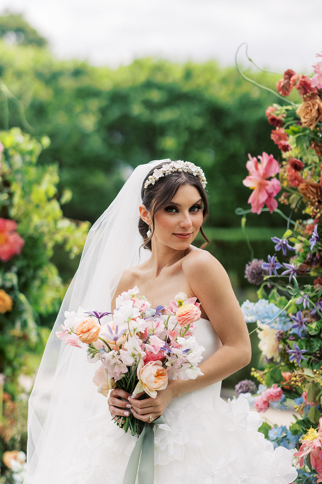 A bride smiles over her shoulder while holding her bouquet around matching colorful flowers in a garden
