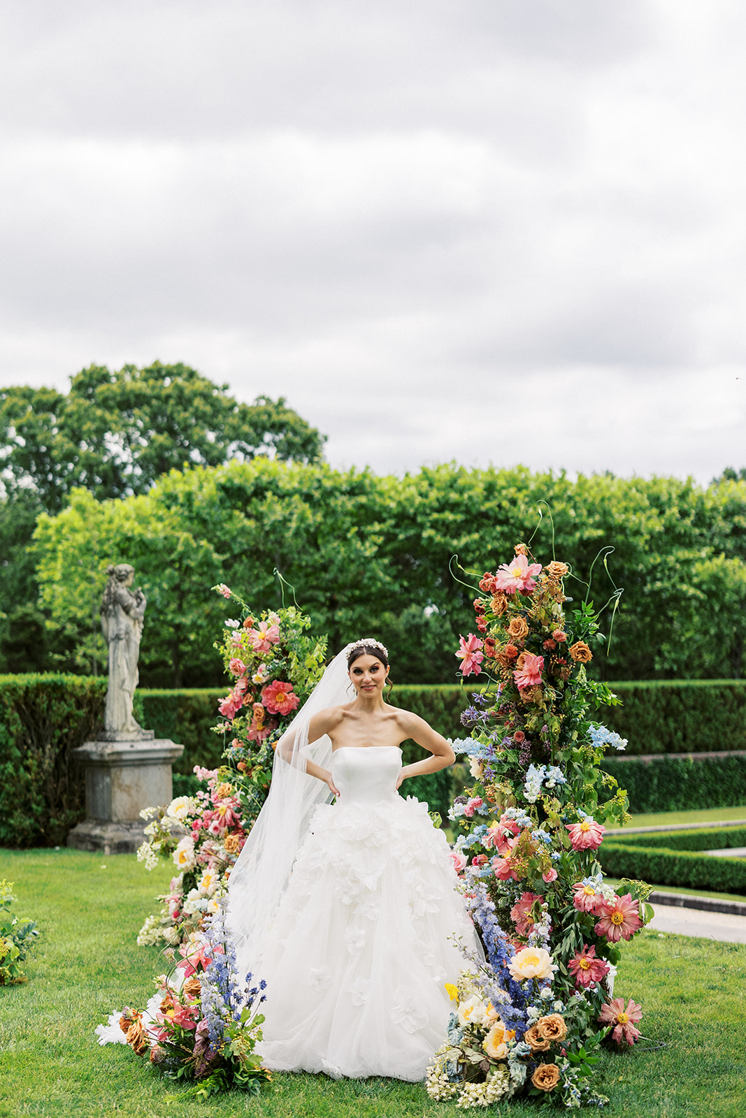 A bride stands amongst a large colorful flower arch with hands on her hips in a garden
