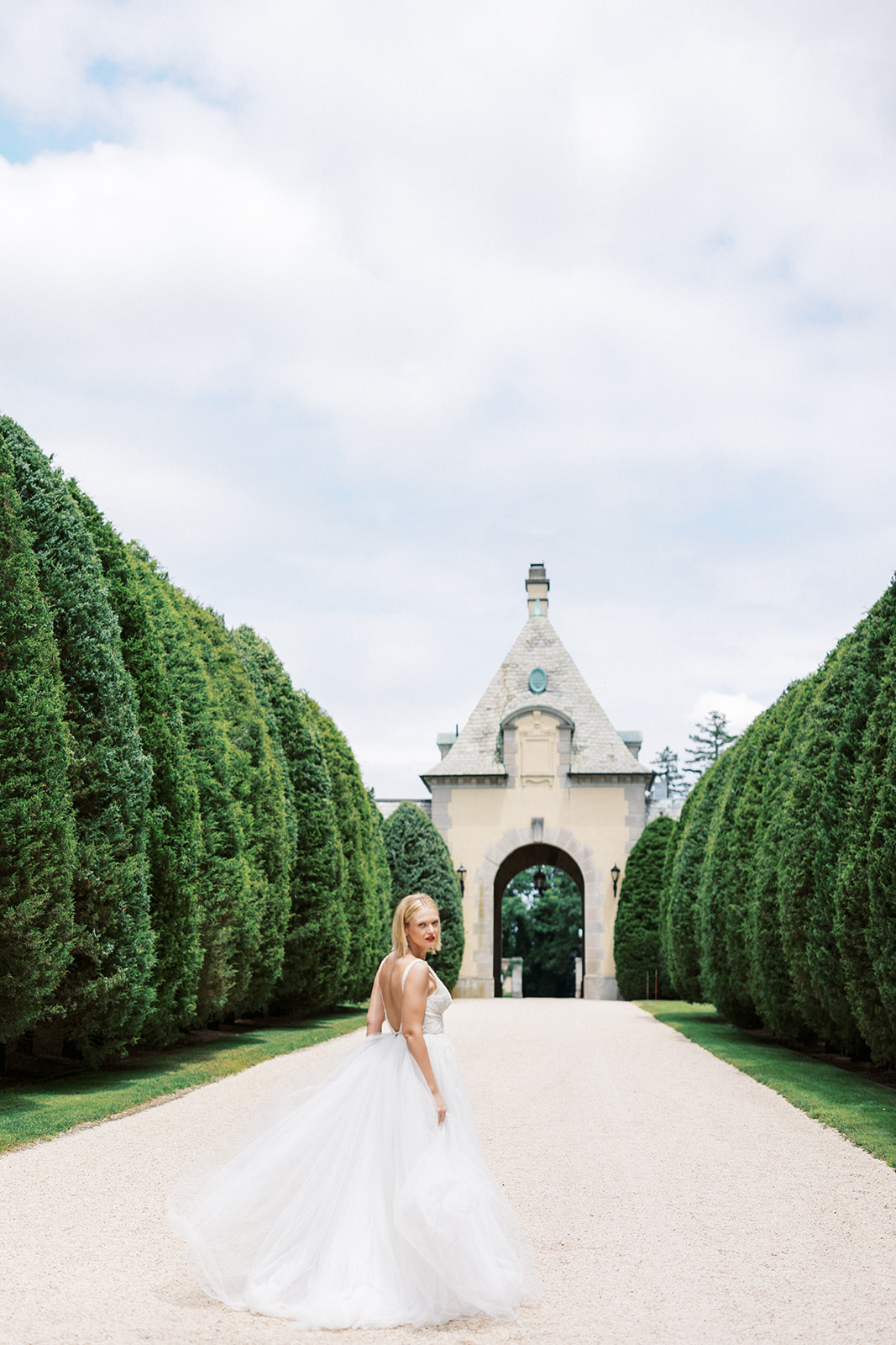 A bride walks down a gravel road lined with tall trees looking over her shoulder