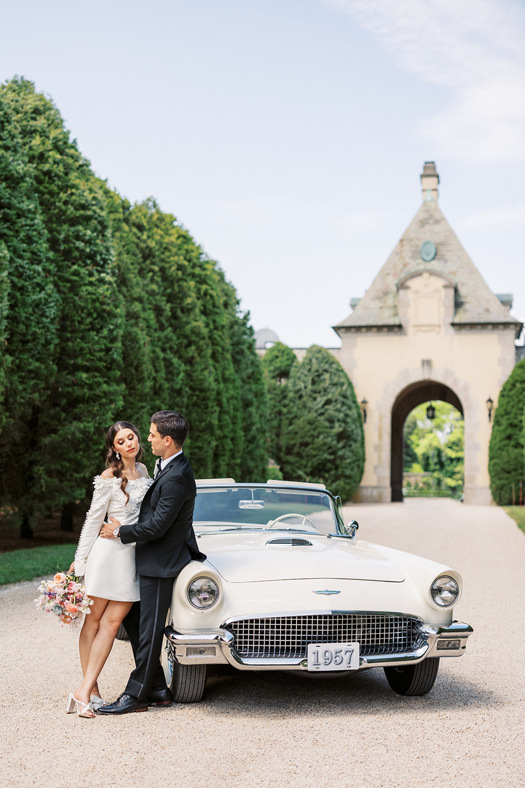 Newlyweds lean on a classes convertible on a tree lined gravel road at the entrance to the Oheka Castle