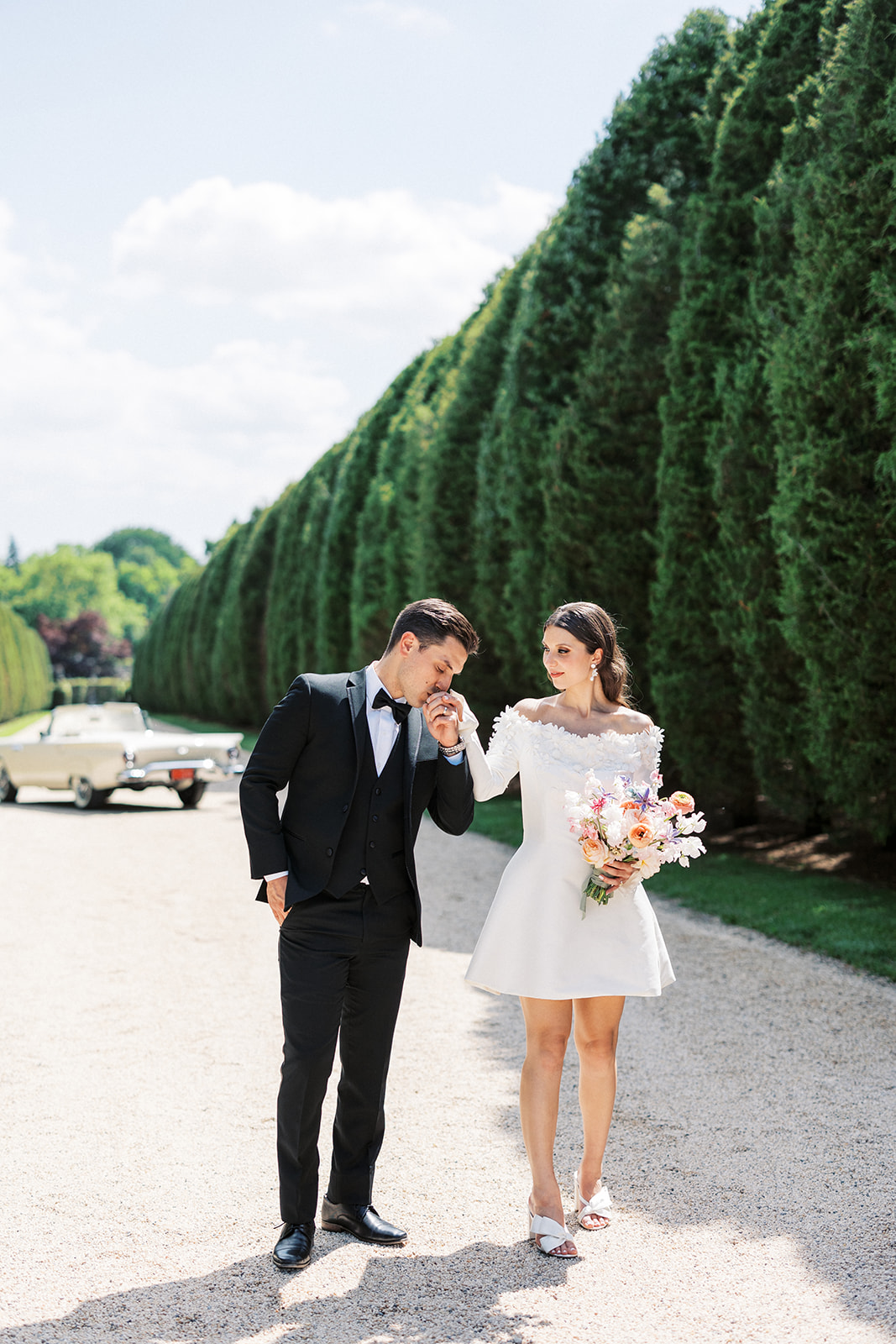 A groom in a black tuxedo kisses the hand of his bride as they walk through a gravel road at the Oheka Castle