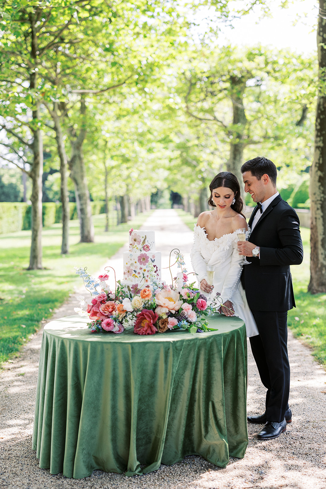 Newlyweds stand at their outdoor cake table holding champagne glasses at an Oheka Castle wedding